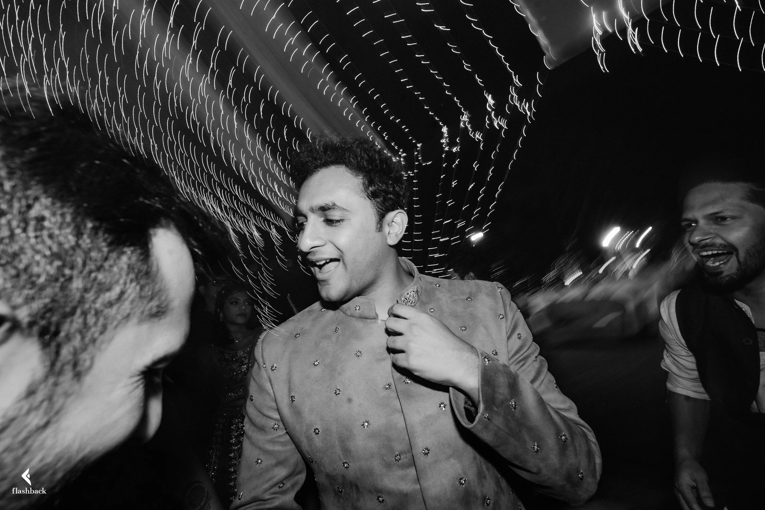 A group of friends celebrating at a nighttime event, with string lights overhead, in black and white. The central figure is a young man wearing a traditional Indian outfit, smiling and engaging with others.