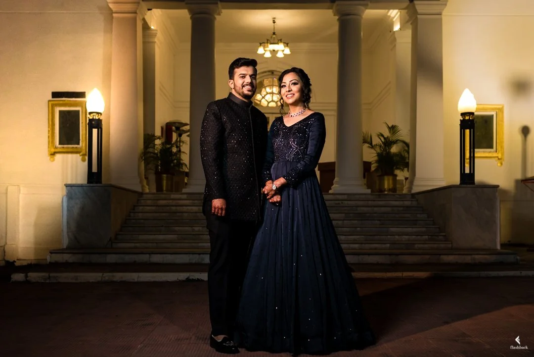 A man and woman dressed in formal attire, standing on outdoor steps in front of a grand entrance with columns and lights, smiling at the camera.