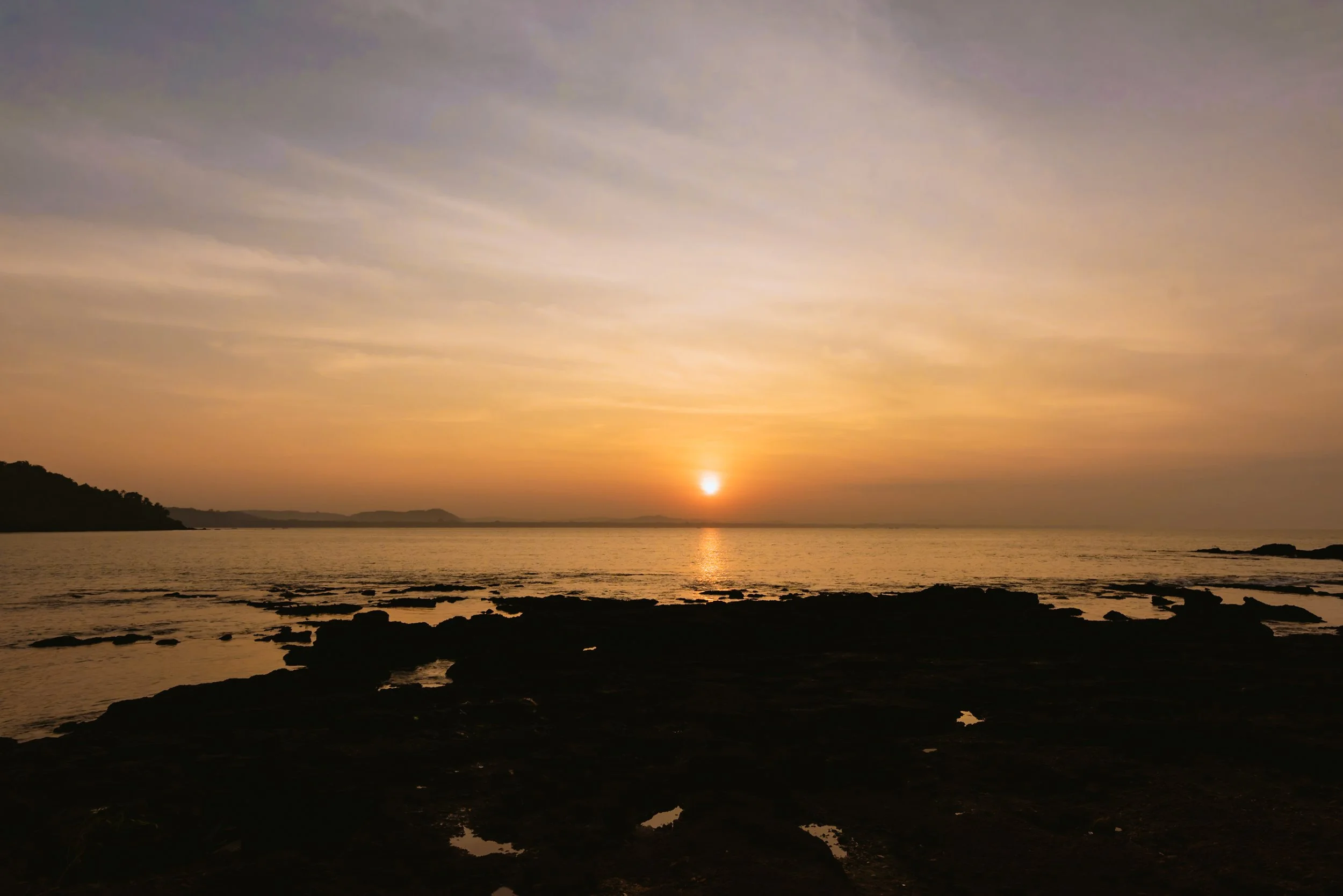 Sunset over a calm body of water with rocky shoreline and a distant landmass on the horizon.