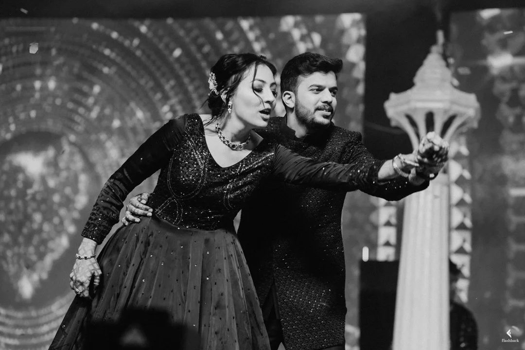 A black and white photo of a woman and man dressed in formal attire, with the woman wearing a flowing dress and the man wearing a glittery jacket, taking a selfie on stage with a backdrop of lights and column decorations.