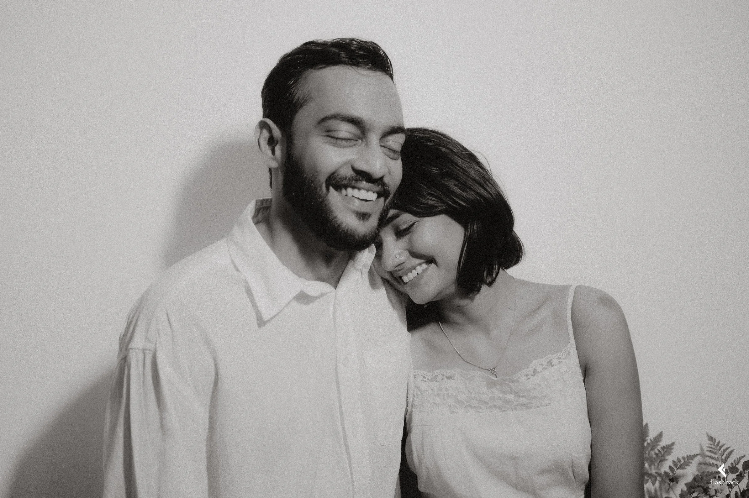 A black-and-white photo of a smiling man and woman with their eyes closed, leaning on each other against a plain wall, with some decorative plants in the corner.