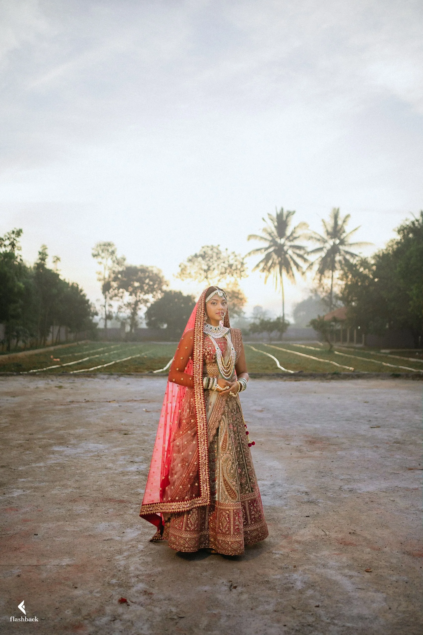 A woman dressed in traditional Indian bridal attire standing outdoors at sunrise or sunset.