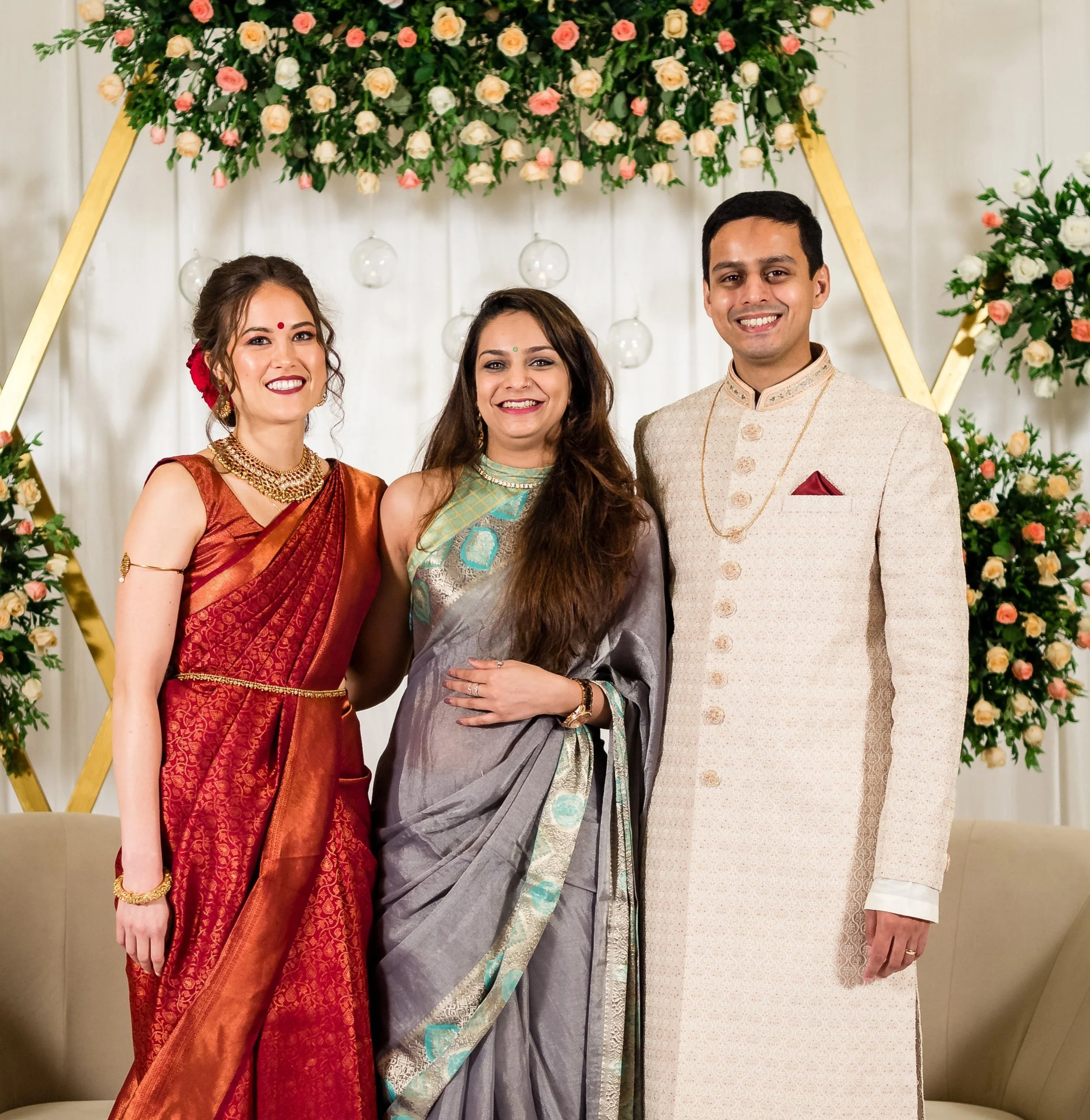 Three people dressed in traditional Indian attire standing in front of floral decorations, smiling at the camera.