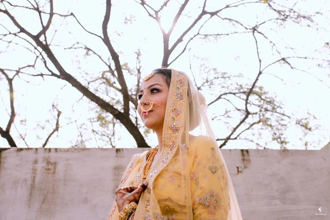 A woman dressed in traditional Indian bridal attire, wearing a yellow and gold embroidered outfit with jewelry, standing outdoors with a bare tree in the background.