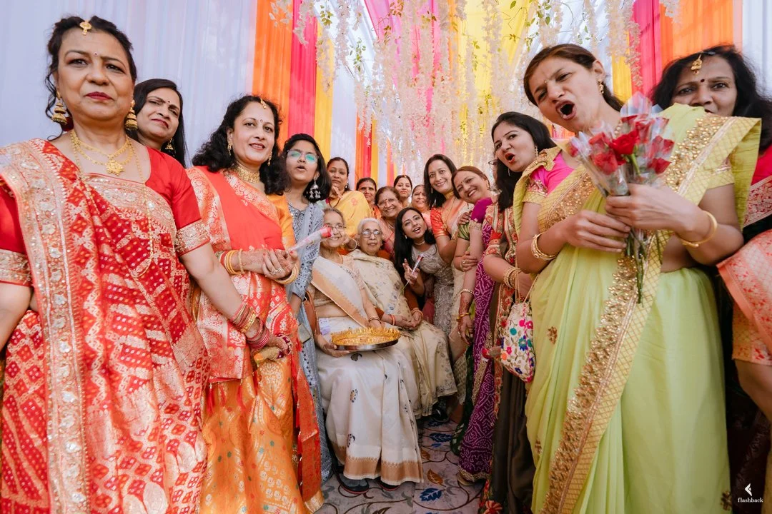 A group of women in colorful traditional Indian sarees gathered around an elderly woman sitting on a chair during a festive celebration. One woman on the right is holding a bouquet of roses and speaking.