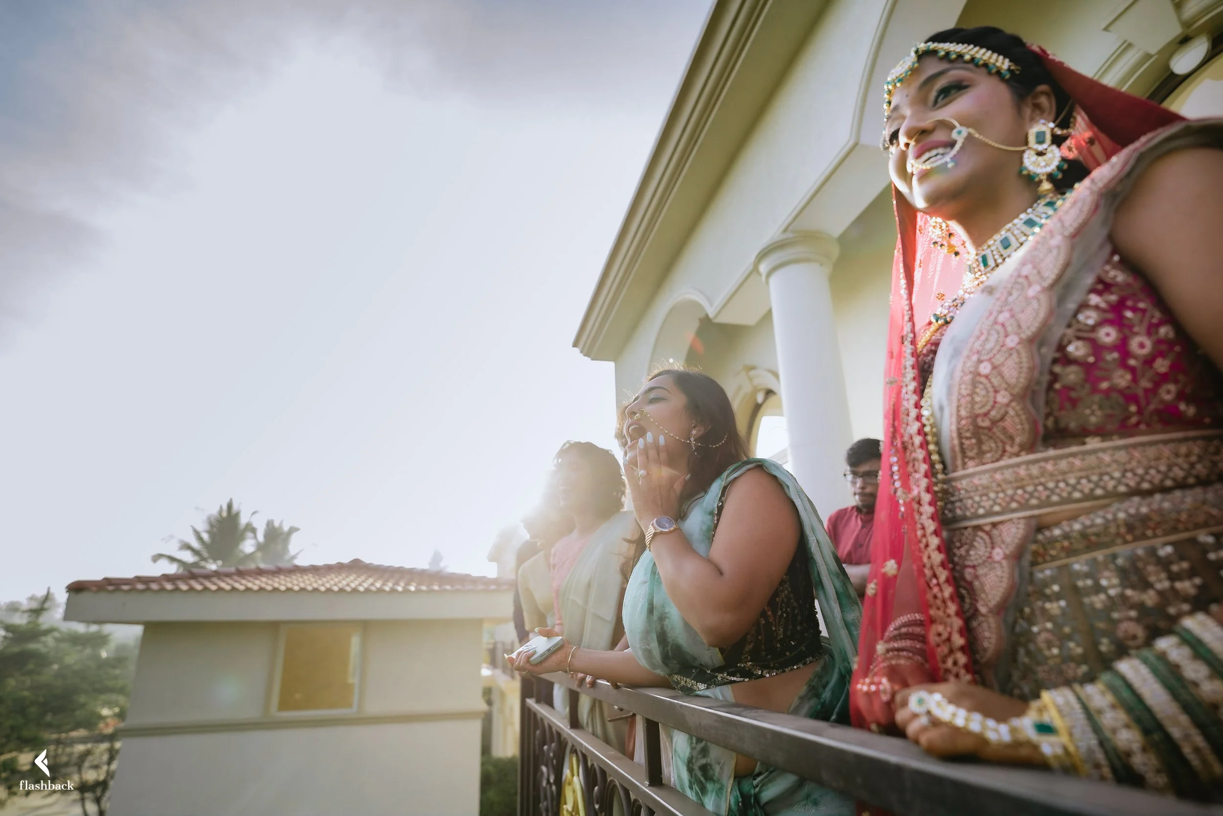 Women dressed in traditional Indian attire, celebrating outdoors on a balcony, smiling and enjoying a festive moment.