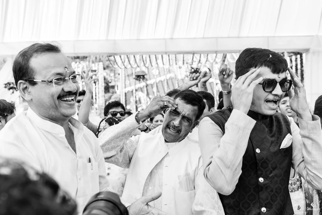 Black and white photo of a group of men in a crowd, some smiling, some with their hands on their heads, in an outdoor setting with decorative elements in the background.