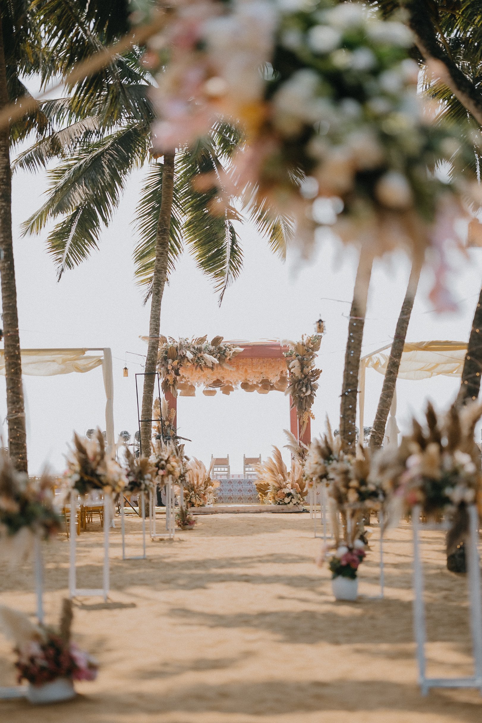 A decorated wedding ceremony setup on a beach with an arch decorated with flowers, palm trees, chairs covered with fabric, and floral arrangements along the aisle.