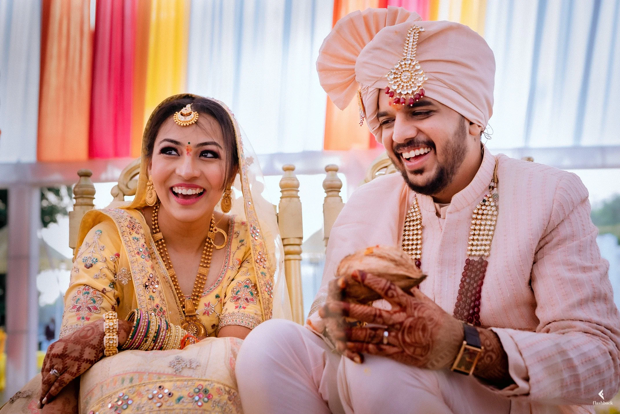 A bride and groom in traditional Indian wedding attire, sitting and smiling during their ceremony, with colorful drapes in the background.