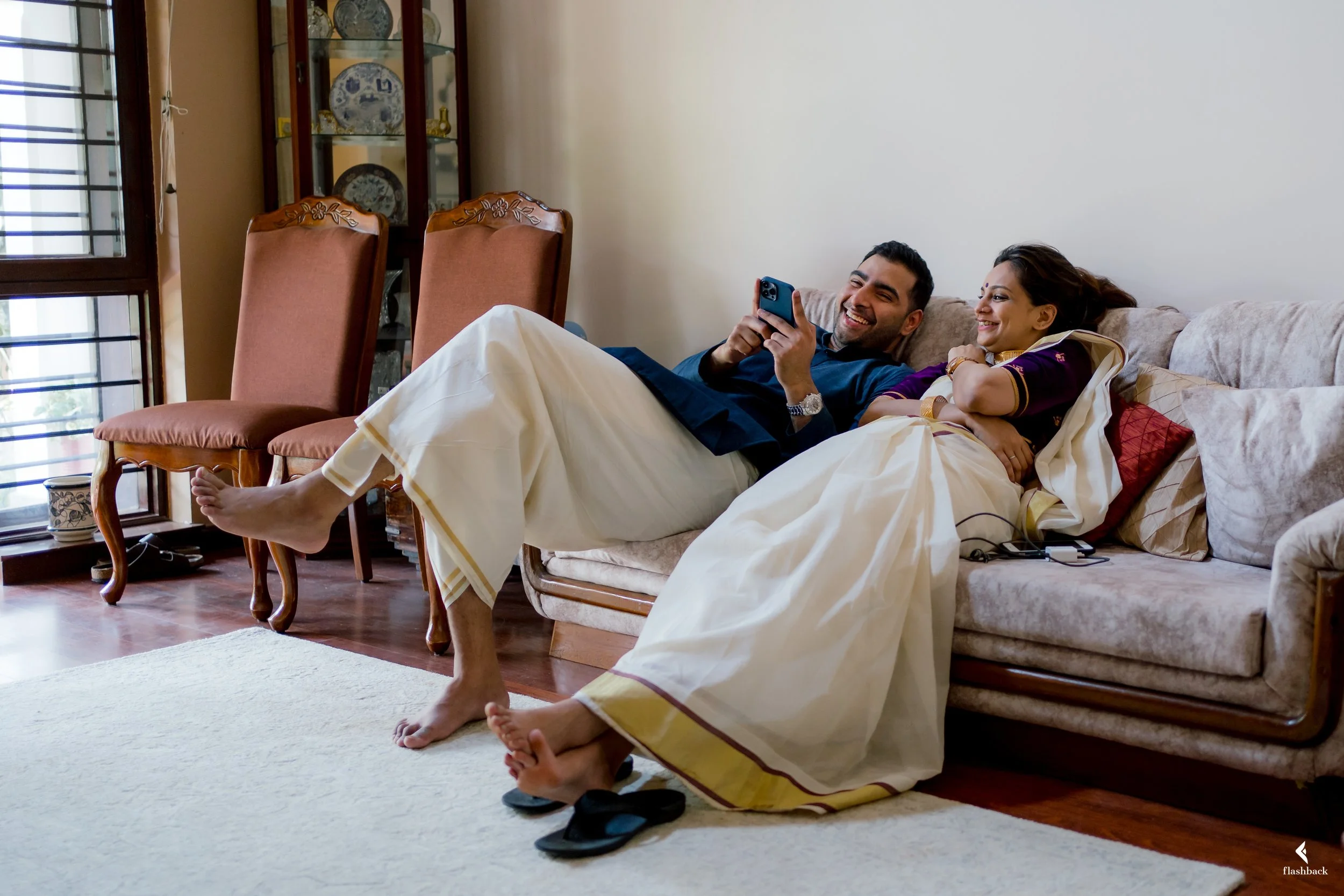 A happy couple lying on a sofa in a living room, looking at a smartphone. The man is wearing a dark blue shirt, and the woman is in traditional Indian attire. They are smiling and relaxed, with some slippers and electronic devices nearby.