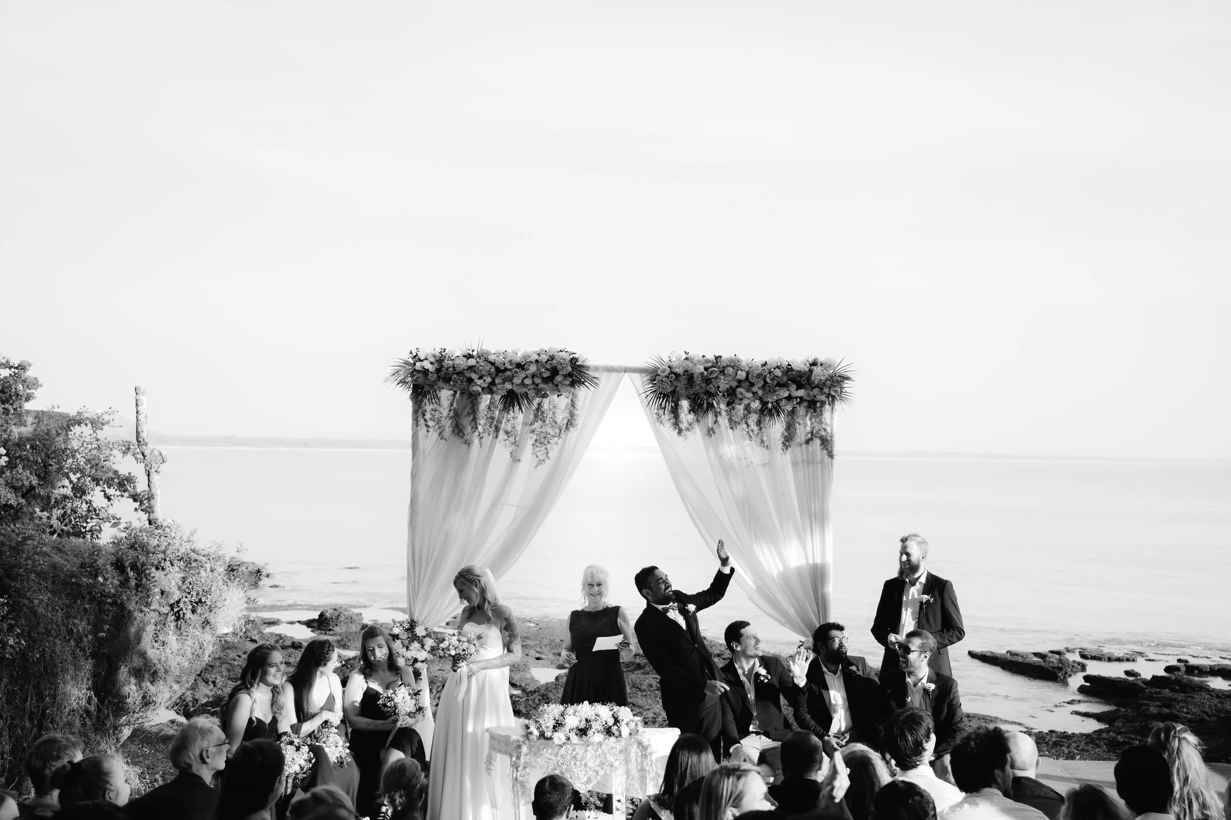 A wedding ceremony taking place on a beach with an outdoor altar decorated with flowers and drapes, with the bride and groom, bridesmaids, and groomsmen present, and guests seated in front.