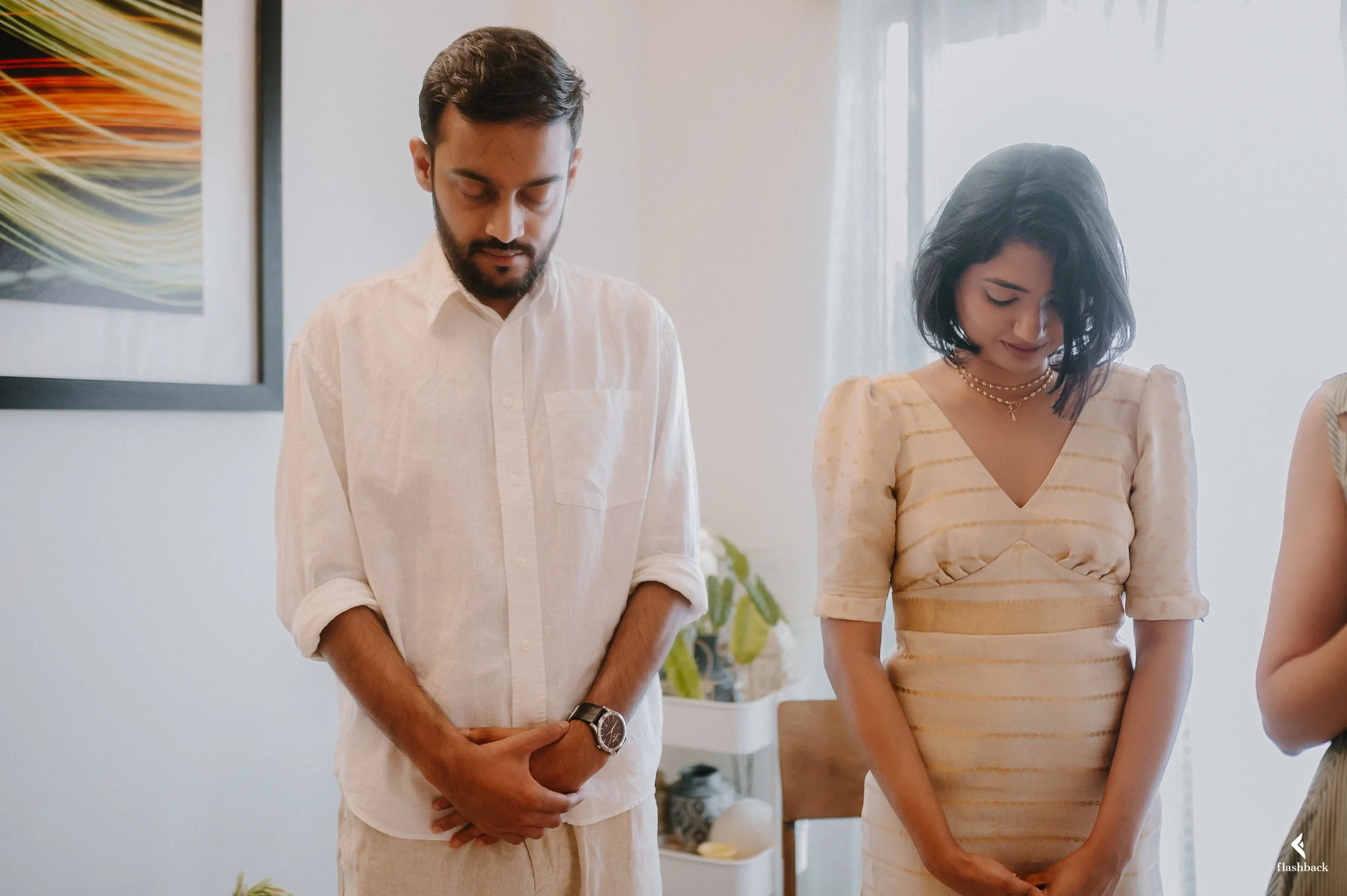 A man and a woman stand with heads bowed and eyes closed in a moment of prayer or reflection inside a well-lit room.