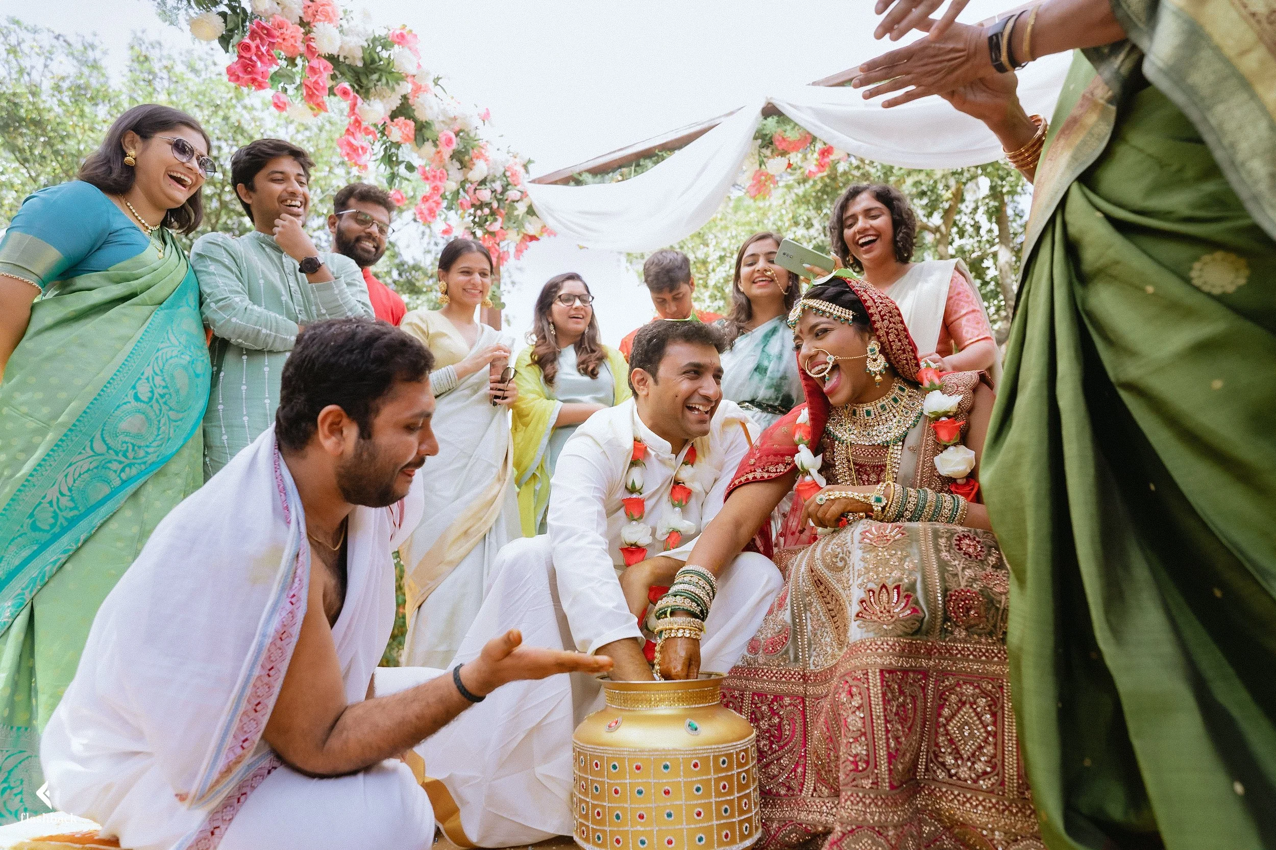 Indian wedding ceremony with the bride and groom participating in a ritual, surrounded by family and friends, outdoor setting with flowers and white drapery.