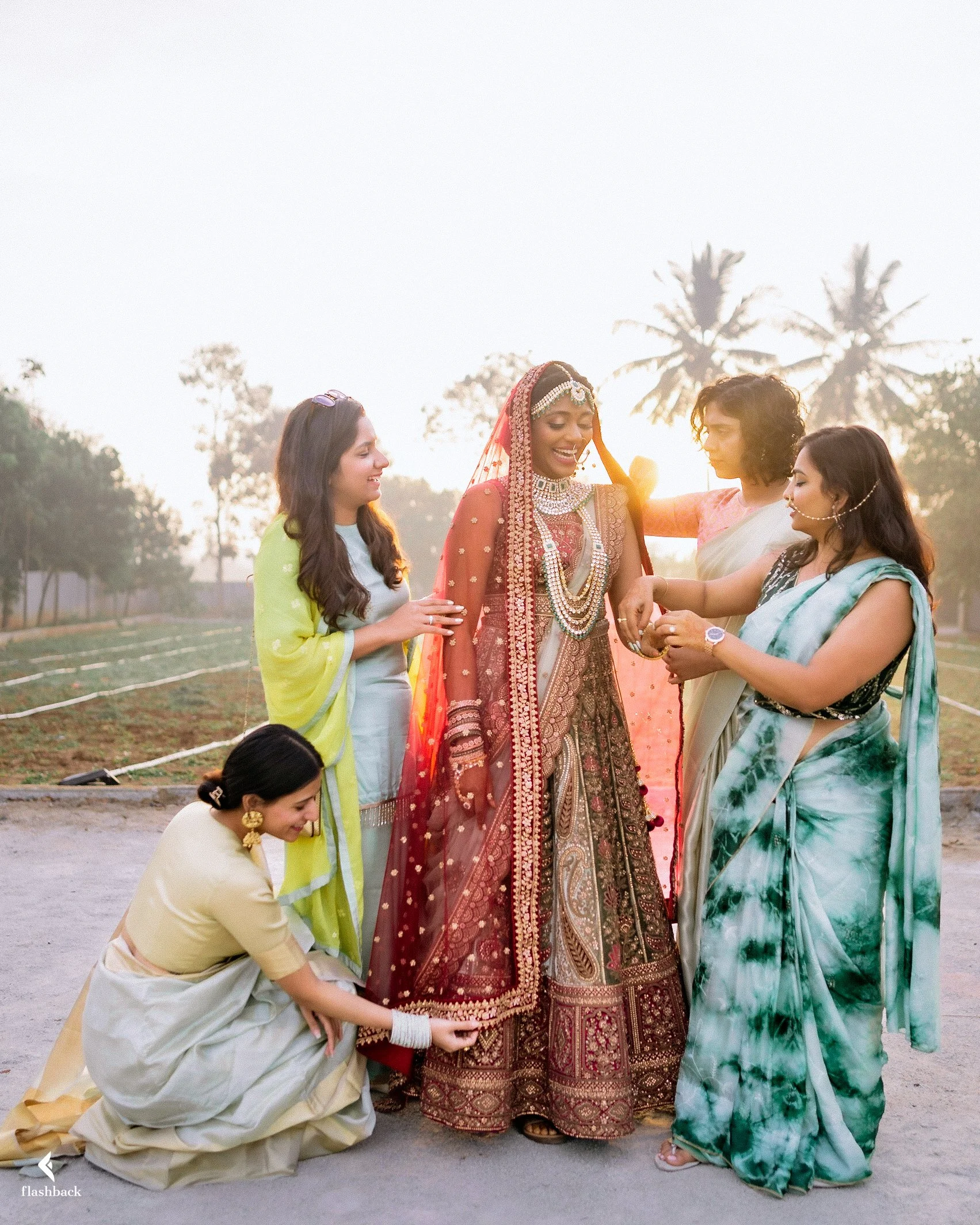 A group of women dressed in colorful traditional Indian attire, including a bride in red and gold, during a wedding ceremony outdoors at sunset.