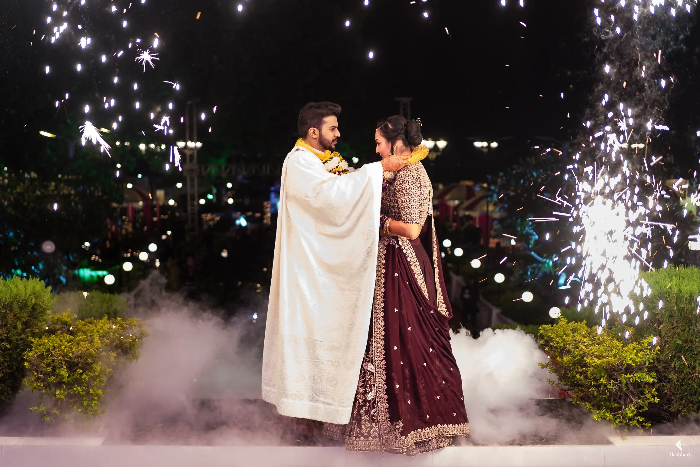 A couple celebrating their wedding at night, with fireworks and smoke effects around them. The man is wearing traditional white outfit and the woman is in a maroon embroidery lehenga.