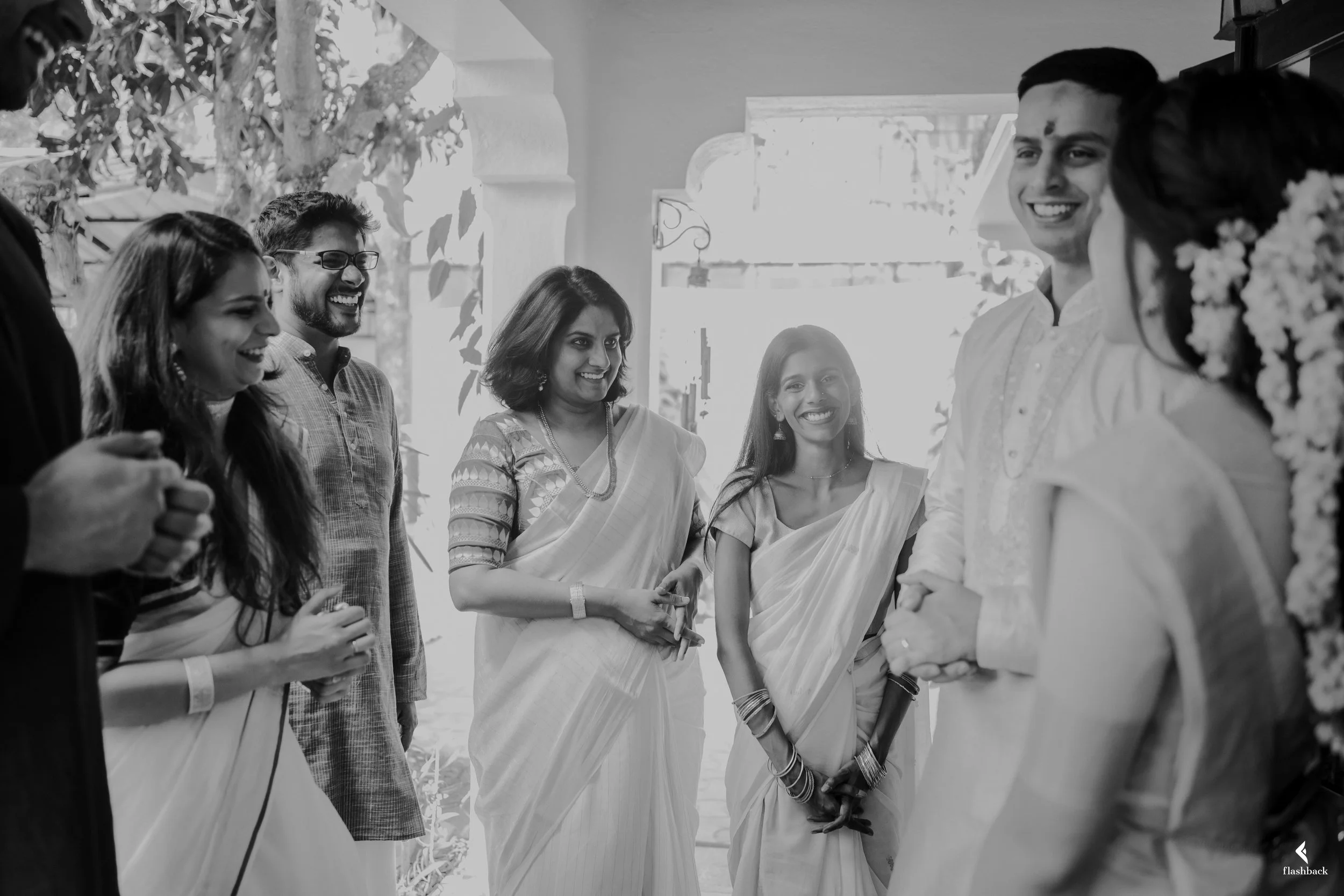 A group of people celebrating, smiling, and dressed in traditional Indian attire, standing outdoors near a white archway with trees in the background.