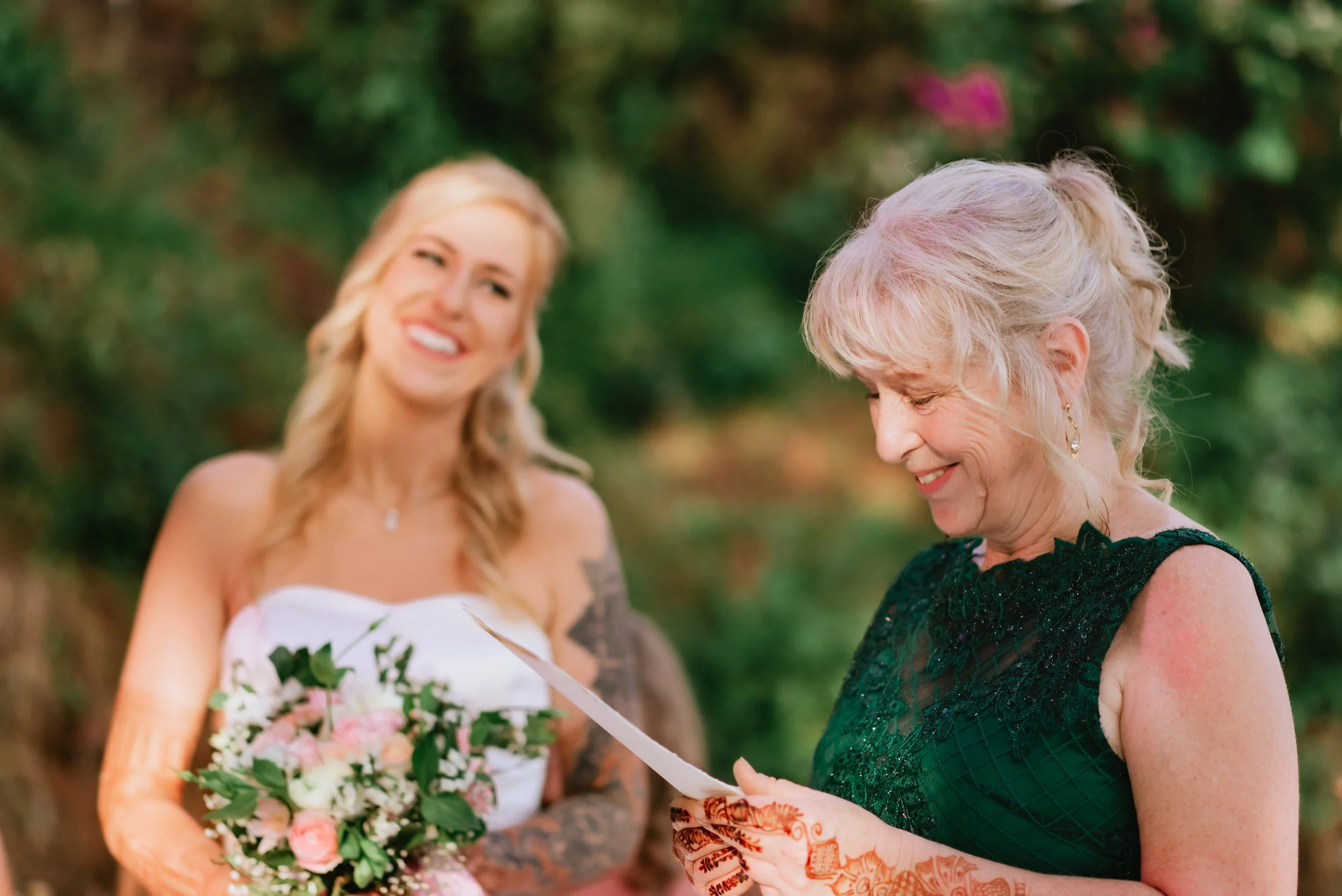 A young woman in a wedding dress is holding a bouquet of pink and white flowers and smiling while an older woman in a dark green dress is reading a letter and smiling outdoors.
