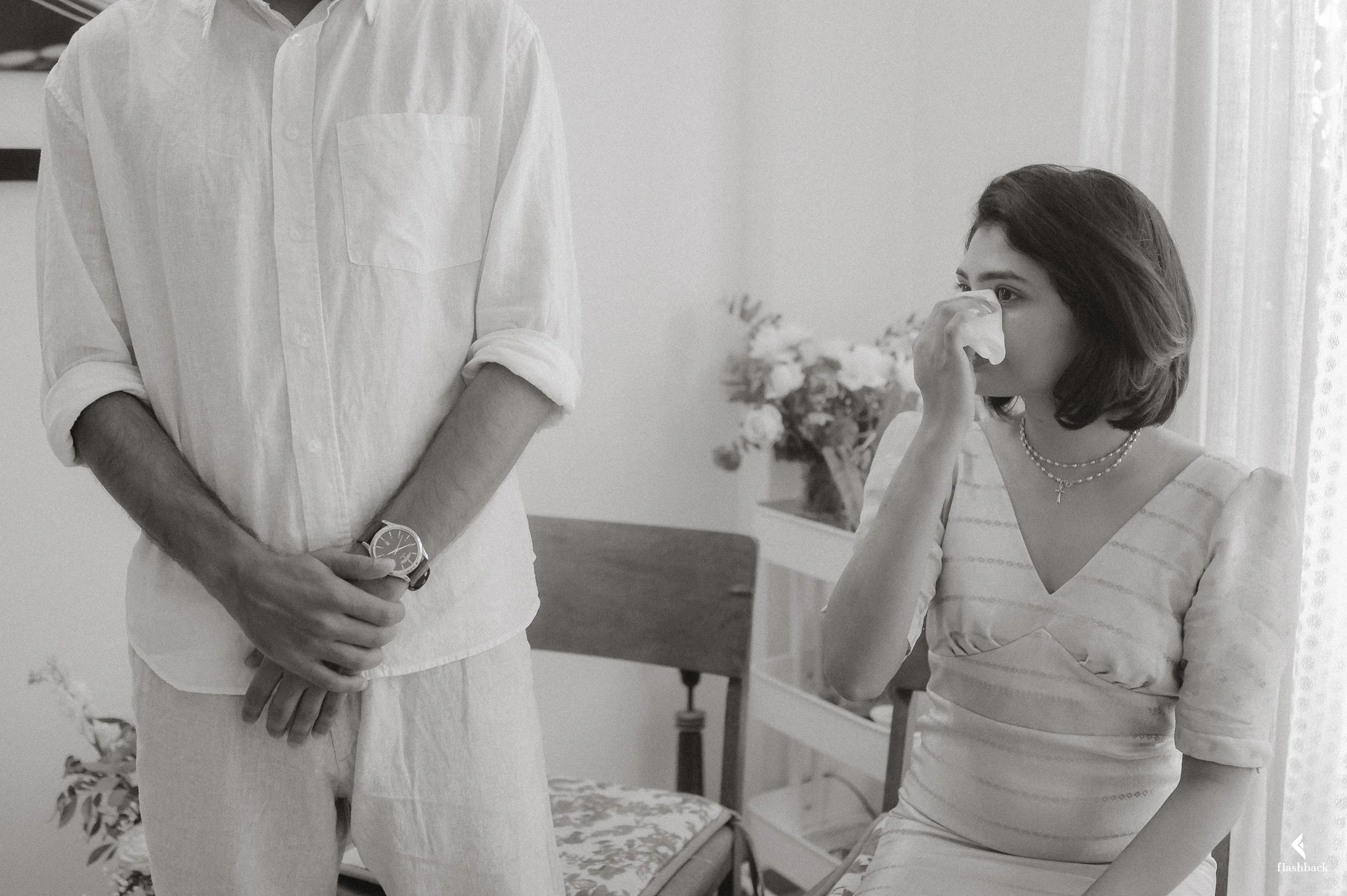 A woman is sitting in a chair, holding a tissue to her nose and crying, while a man stands nearby with his hands clasped. The scene appears emotional and is set in a room with a table, flowers, and curtains.