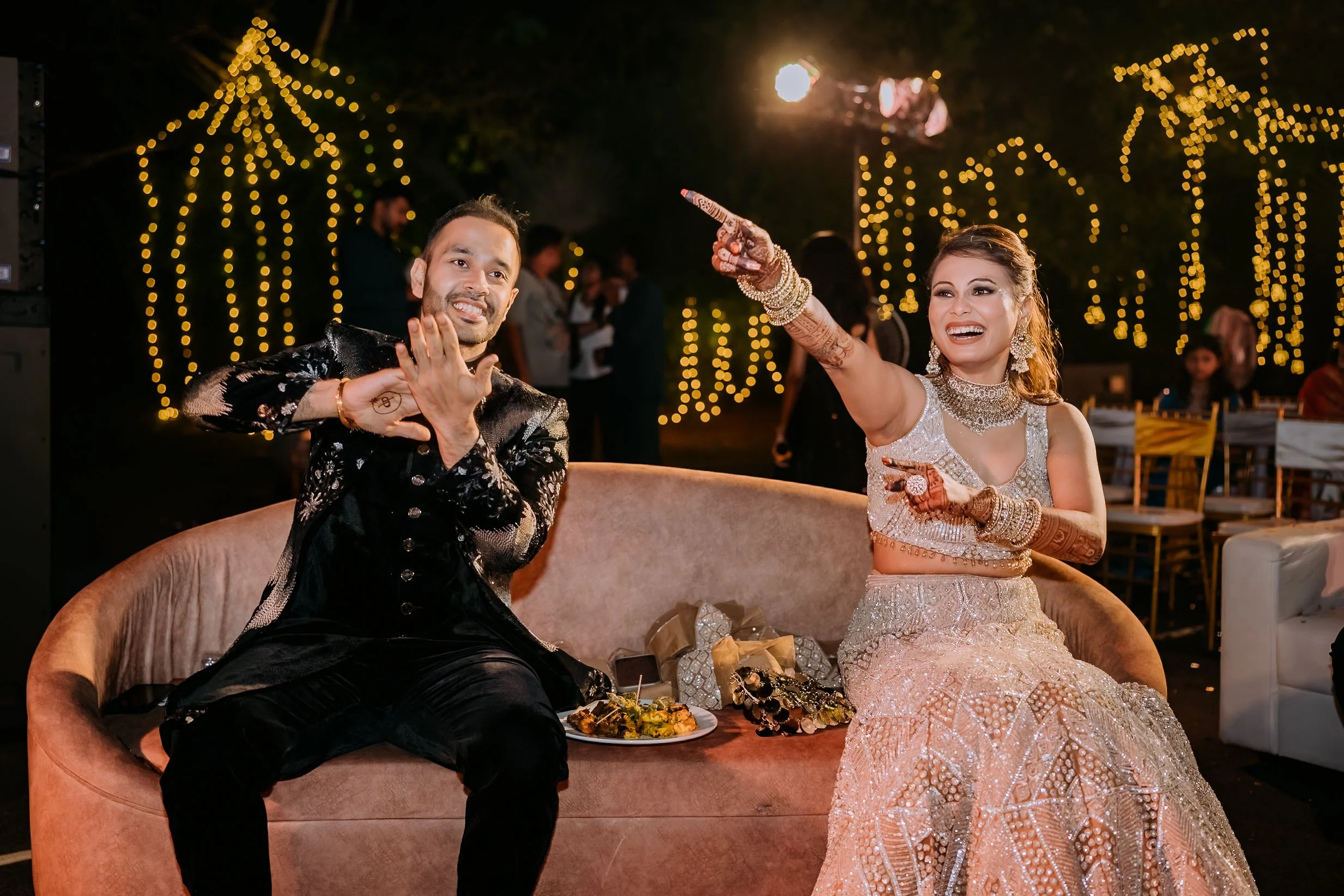 A smiling bride and groom sitting on a sofa at an outdoor wedding reception at night with string lights in the background. The bride is wearing a decorated traditional dress and jewelry, while the groom is dressed in a black embroidered sherwani. The