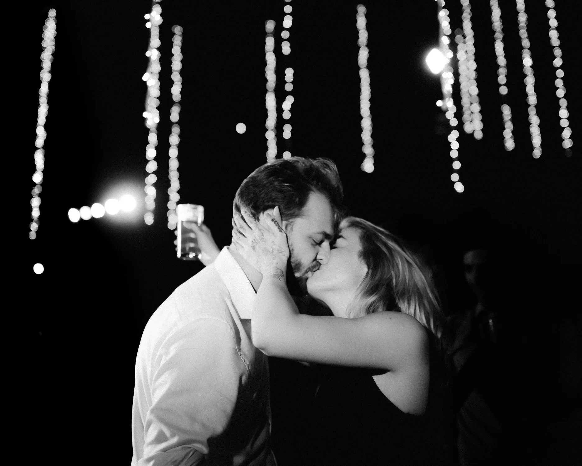 A couple sharing a kiss at night with string lights in the background, in black and white.