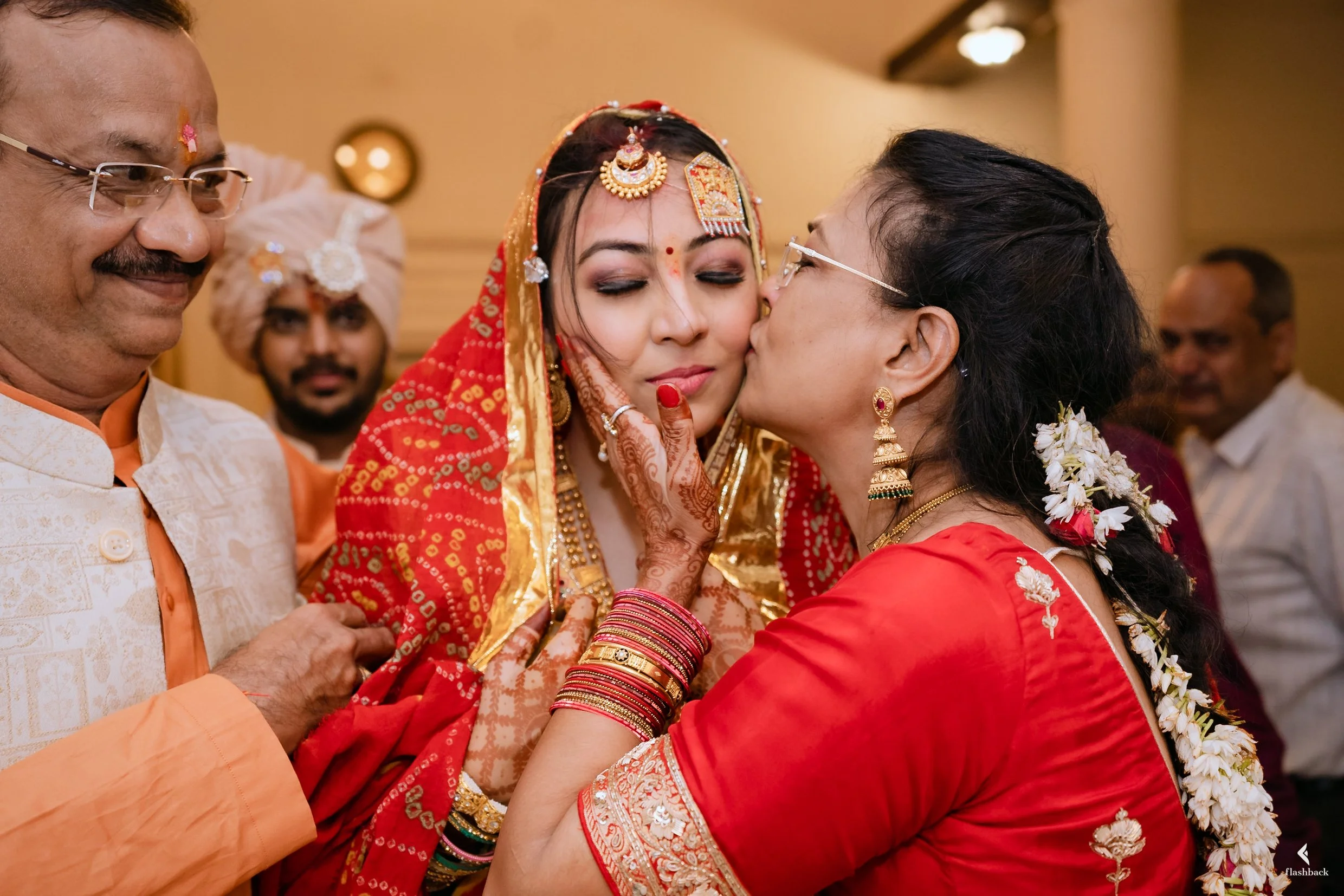 An Indian bride dressed in traditional red and gold attire with intricate jewelry, being kissed on the cheek by an older woman wearing a red saree and flowers in her hair during a wedding celebration. Other guests are visible in the background.