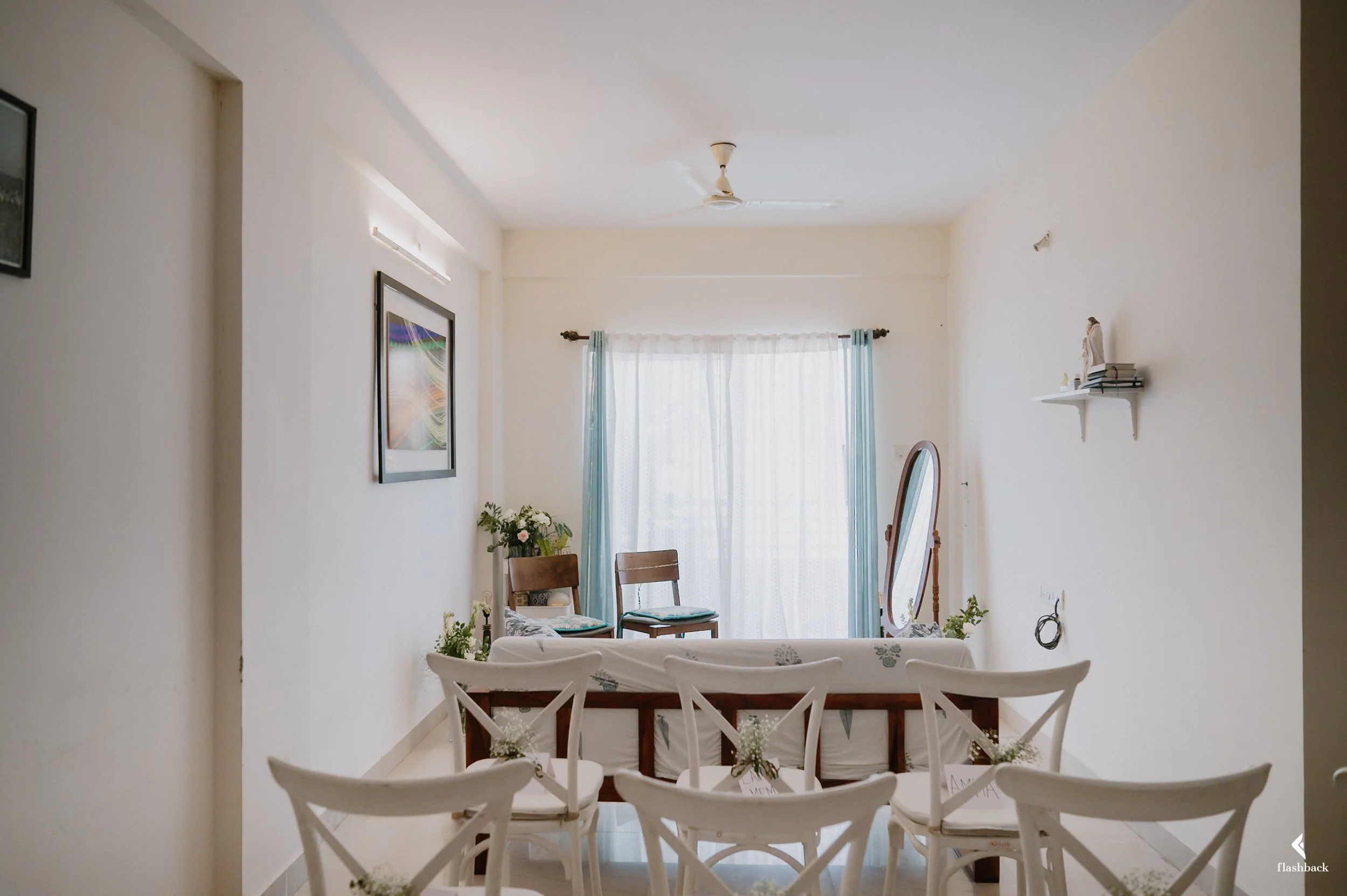 Empty living room with white walls, white chairs decorated with small flowers, a wooden bench, two chairs near a window with blue curtains, a standing mirror, and a small shelf with books and figurines.