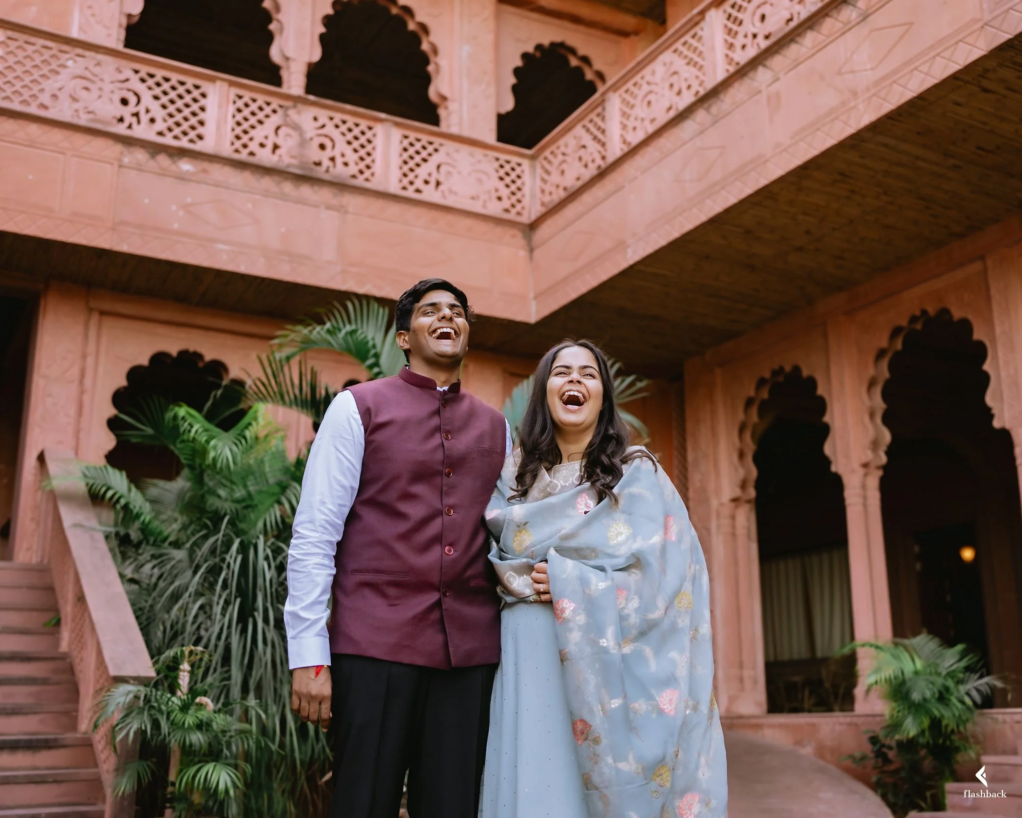 Two smiling people, a man and a woman, standing together in front of an ornate pink sandstone building with lush green plants around them. They appear happy and are dressed in traditional Indian attire.