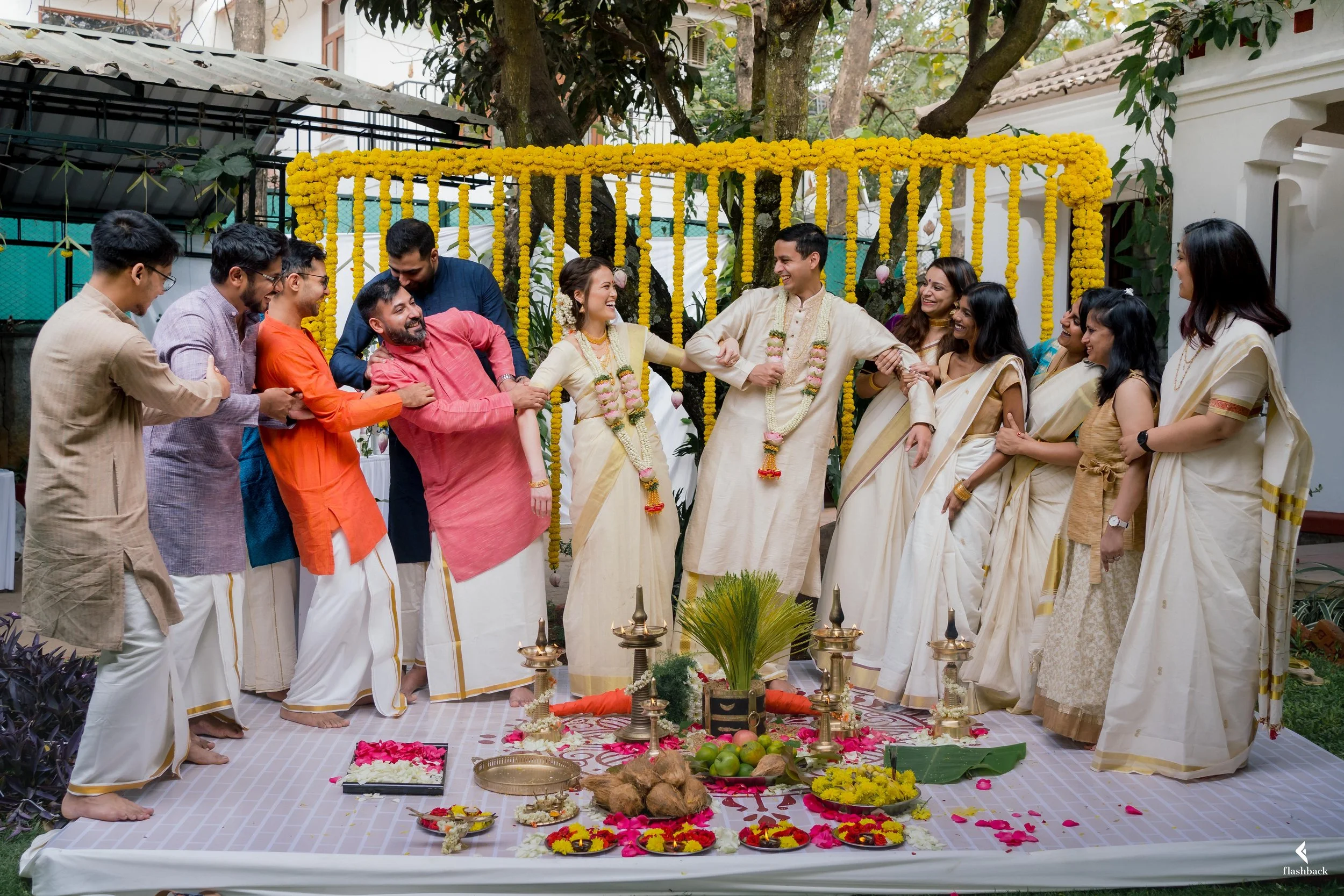 Group of people celebrating a traditional Indian wedding ceremony outdoors, with decorated backdrop and ritual items on the ground.