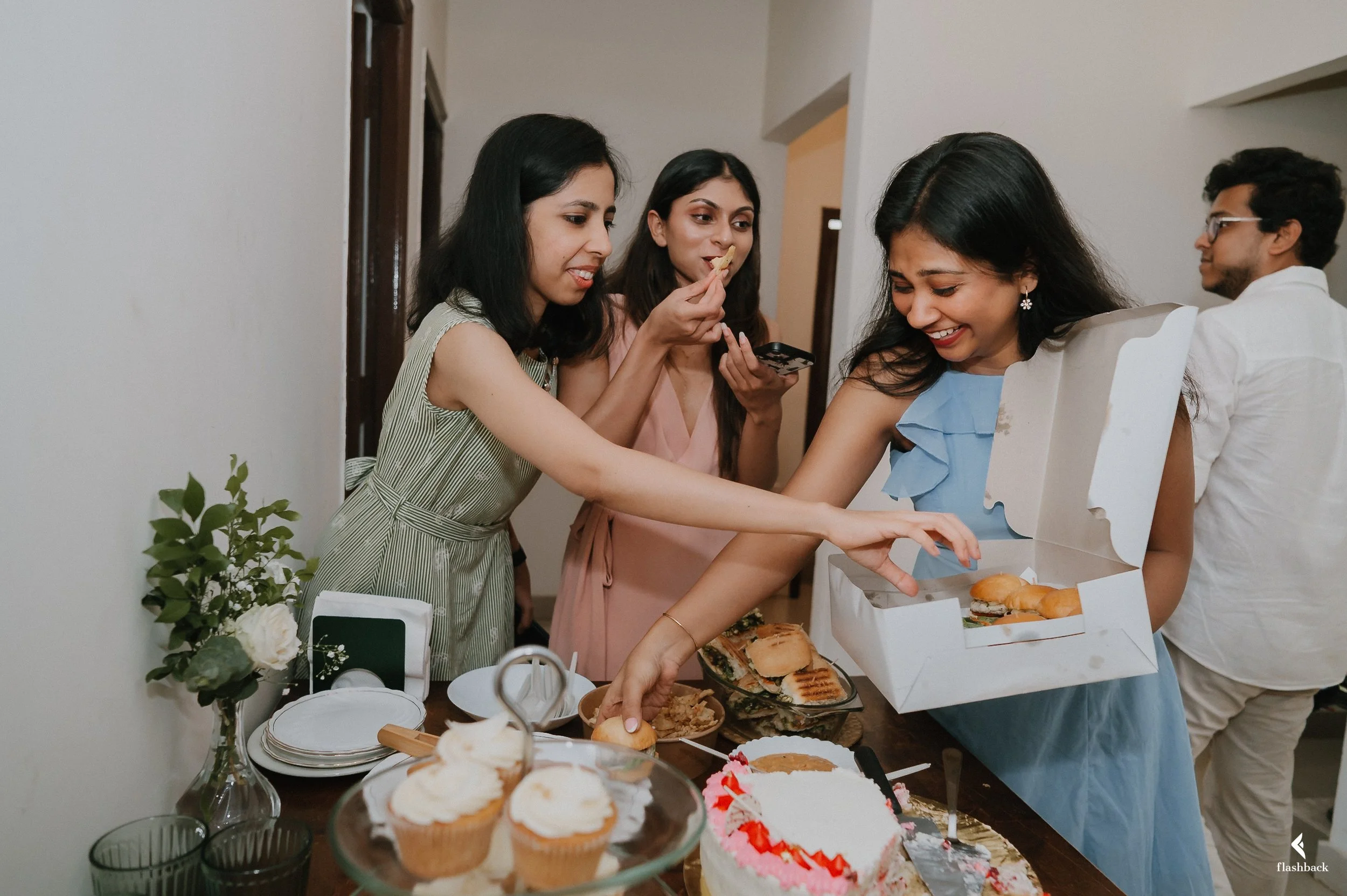 Group of women at a celebration, sharing and serving food, with a table of cupcakes, cake, and snacks.