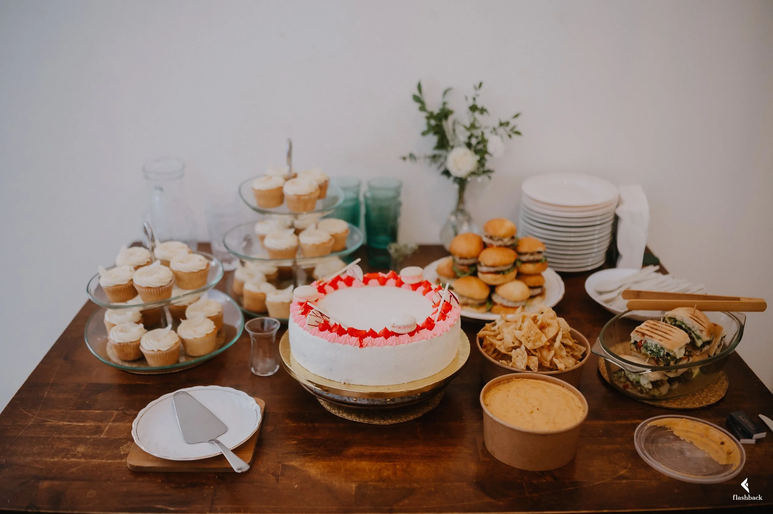 Table with cupcakes, a cake, sliders, chips, dips, and plates, decorated for a celebration.