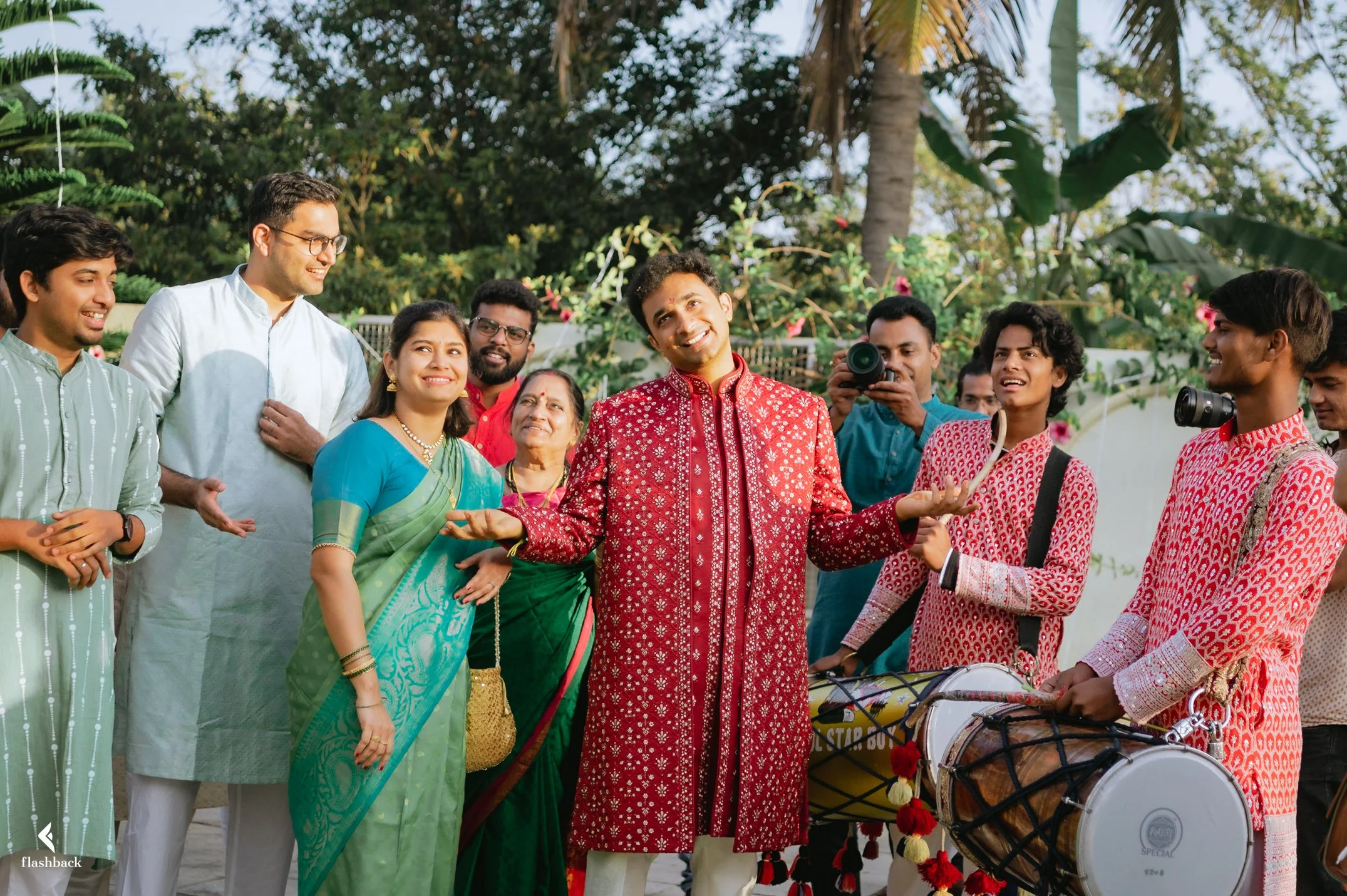 Group of people celebrating outdoors with traditional Indian attire, including a man in a red sherwani and others with musical instruments, smiling and enjoying the occasion.