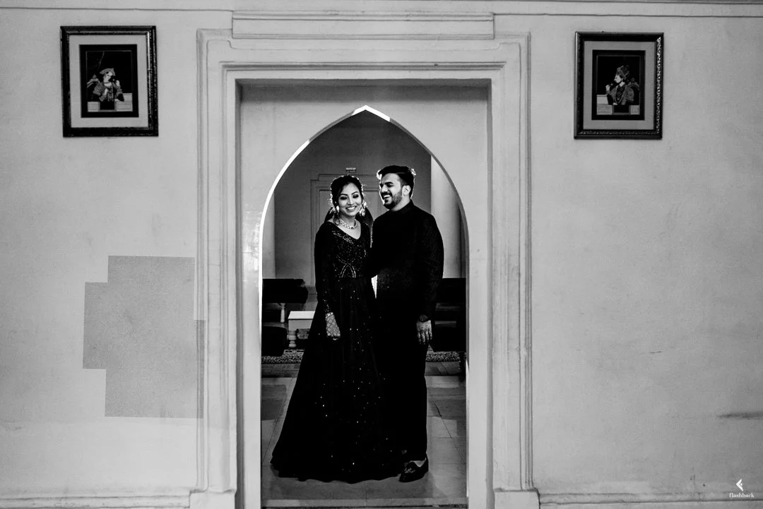 A black and white photo of a smiling couple dressed in formal attire, standing together inside a decorative arched doorway, with framed pictures on the wall behind them.
