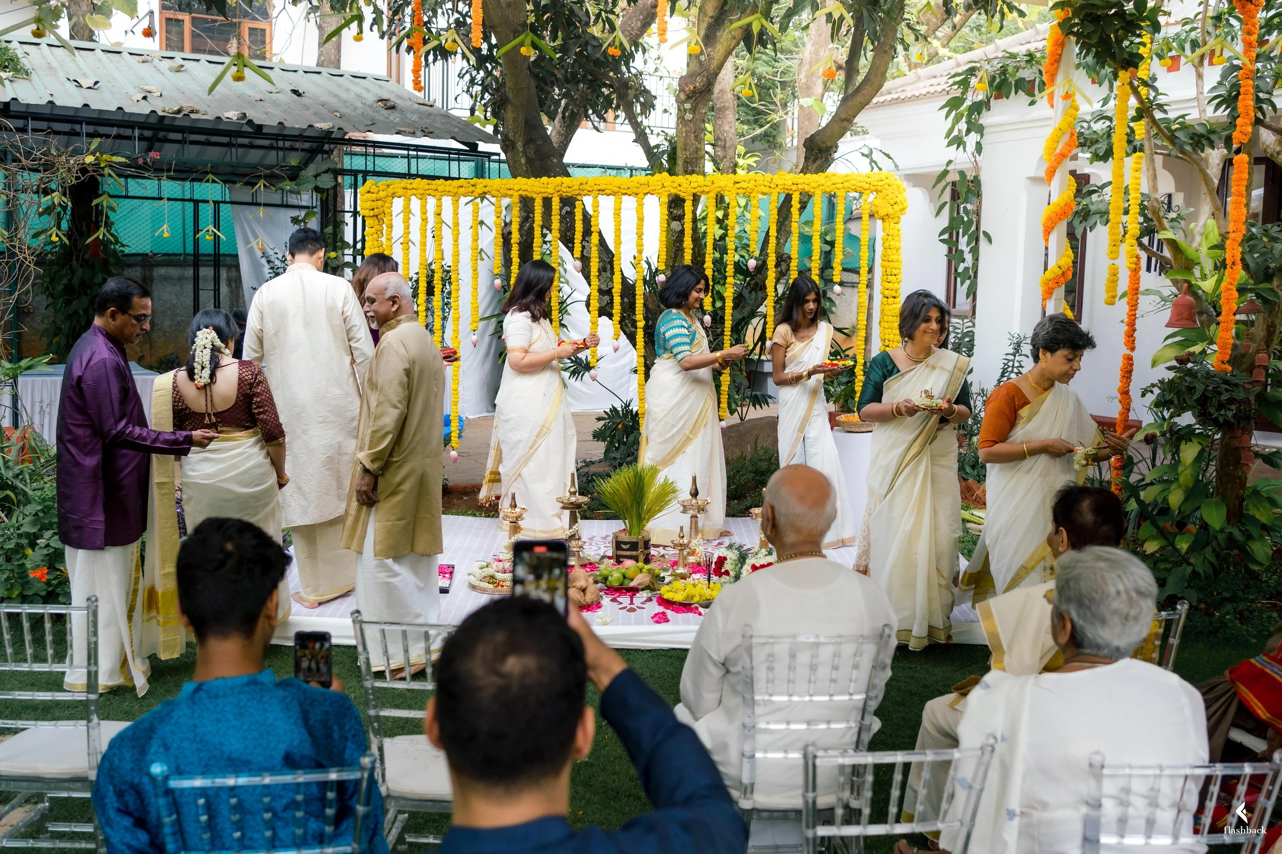 A traditional Hindu wedding ceremony outdoors with women dressed in white sarees and men in traditional attire, decorated with yellow marigold flowers and hanging floral garlands.