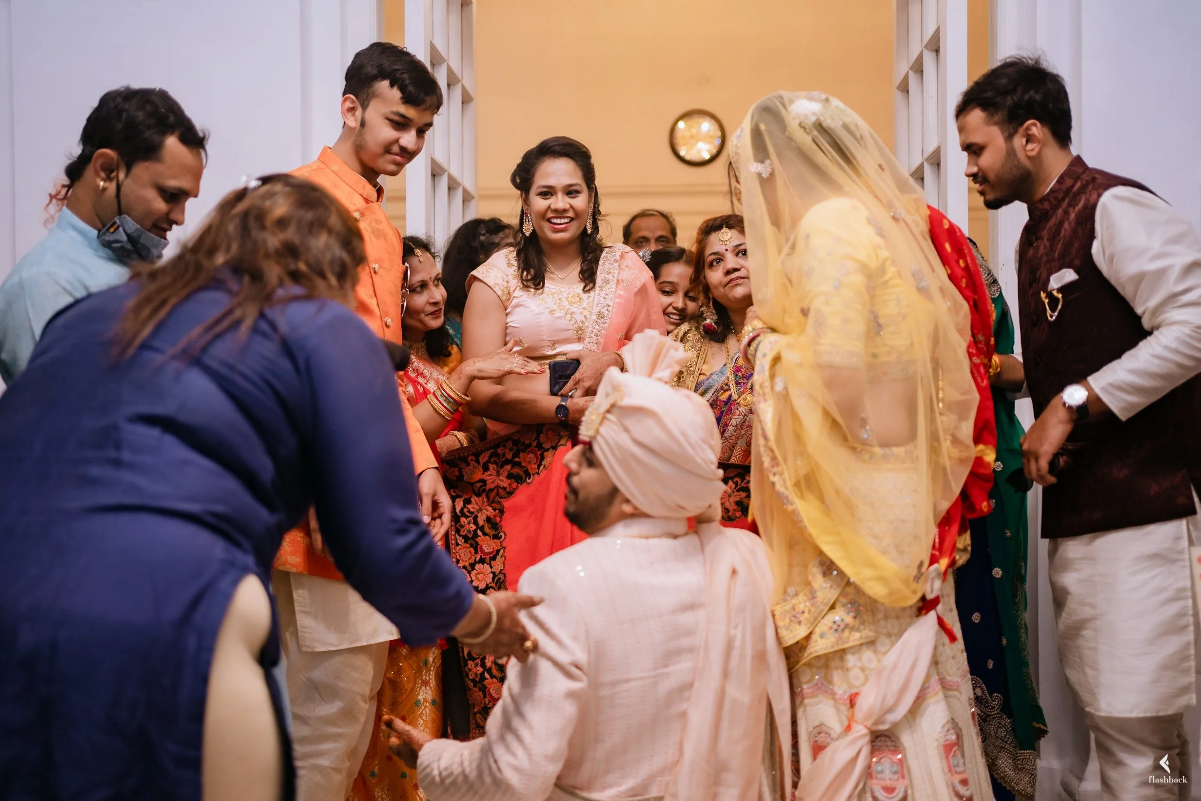 People celebrating an Indian wedding, with the bride wearing a yellow saree and veil, surrounded by family and friends in colorful traditional attire, inside a decorated indoor setting.