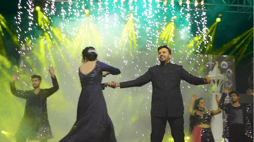 A group of people dancing on a stage with yellow and green lighting, while sparks fall from above, celebrating.