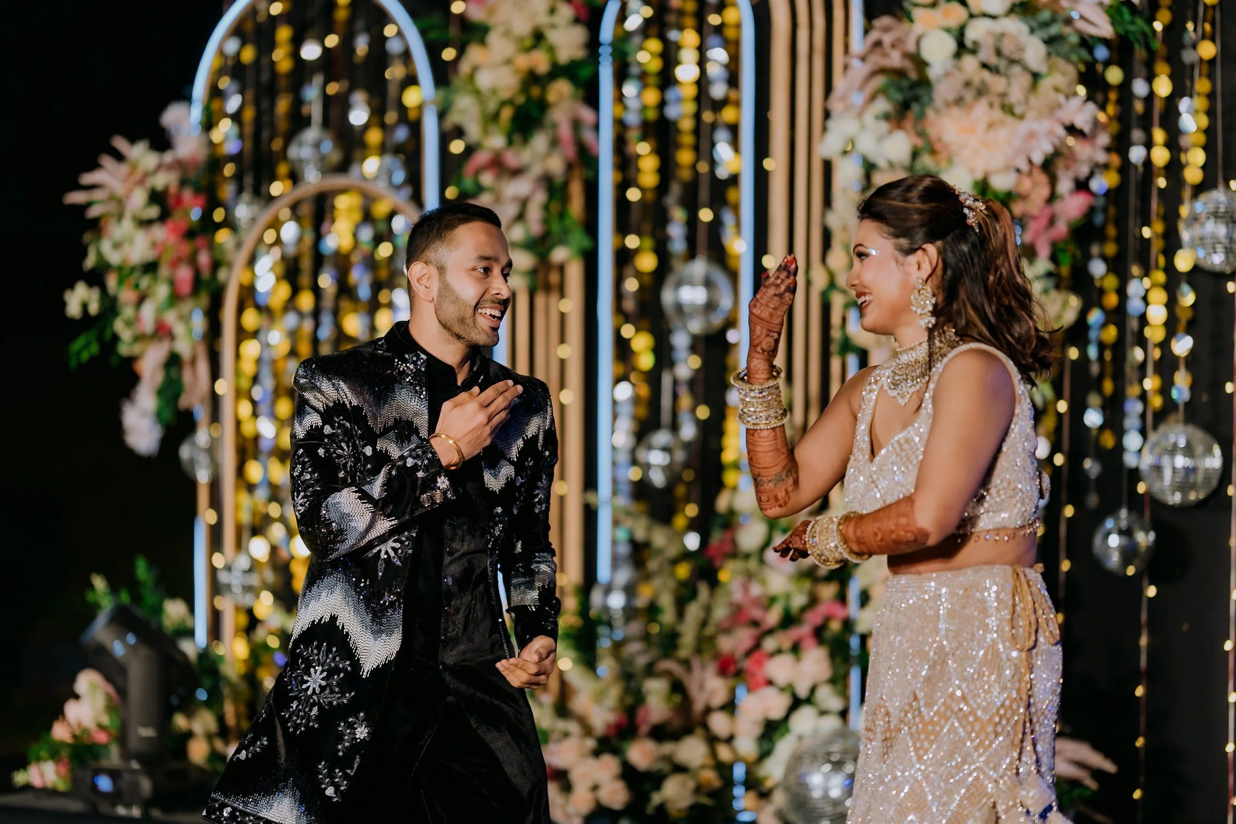 A man and woman sharing a joyful moment at a wedding reception. The man, dressed in a black and silver jacket, is smiling and touching his chest. The woman, in a shimmering traditional dress and jewelry, is smiling and touching her forehead with her 