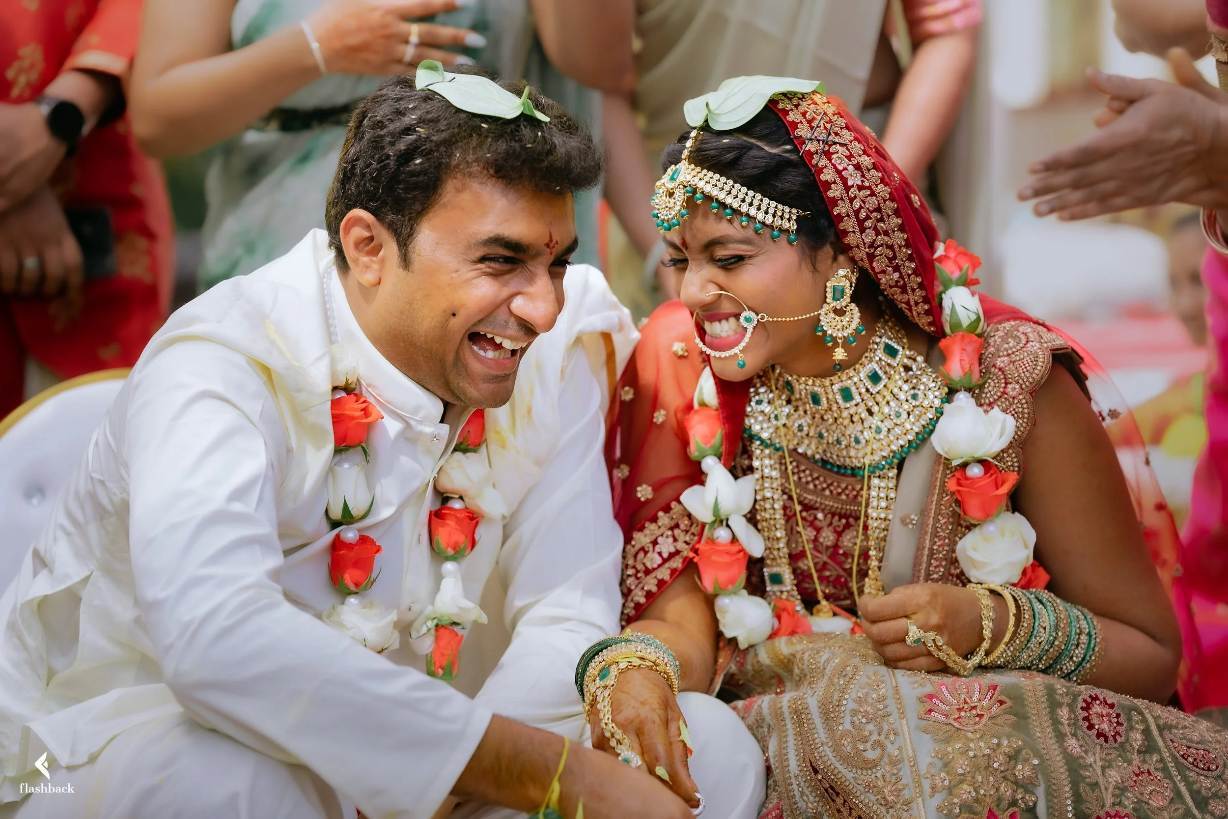 A joyful Indian couple dressed in wedding attire, surrounded by friends and family, celebrating their wedding ceremony. The bride wears traditional jewelry and a red and gold saree, while the groom is in a white outfit. Both are smiling widely, excha