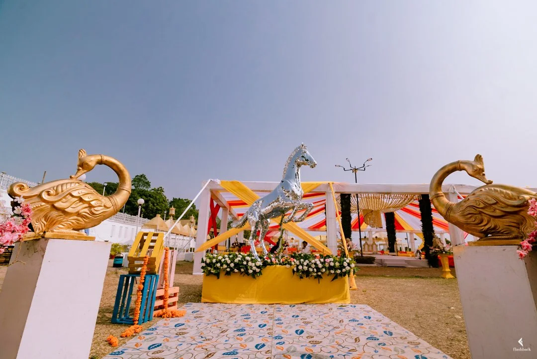 Decorative celebration setup featuring a silver horse statue, two golden swan statues, floral arrangements, and a colorful canopy, likely for an outdoor event or party.