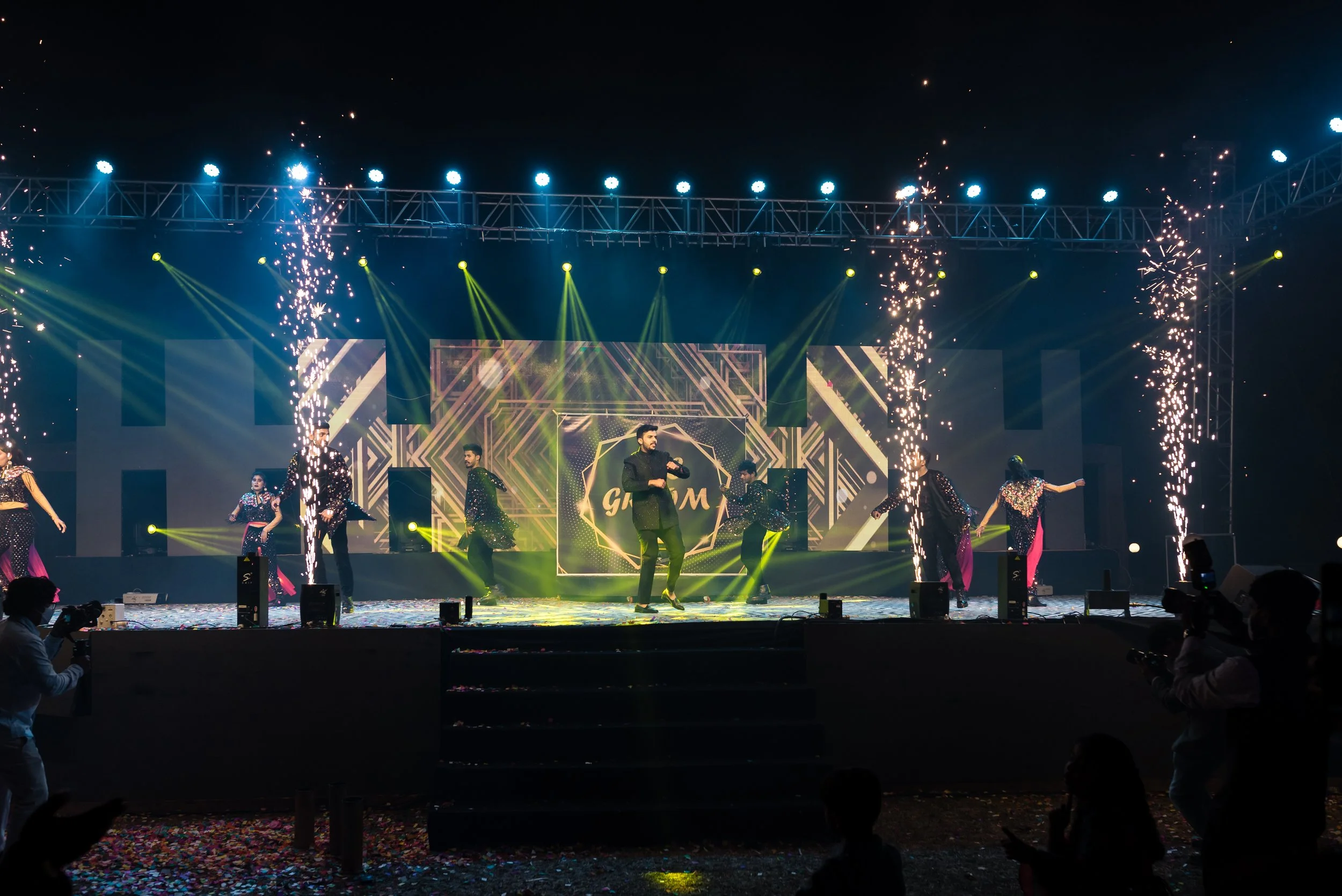 Stage with performers dancing, colorful lights, fireworks, and a digital backdrop displaying the word 'Glam.' Audience members are visible in front and capturing the event with cameras.