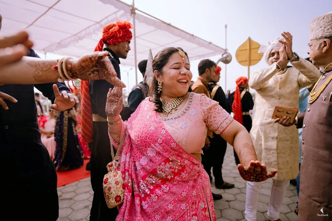 Indian woman in a pink saree dancing at a celebration with men and women around her, some in traditional attire, at an outdoor event.