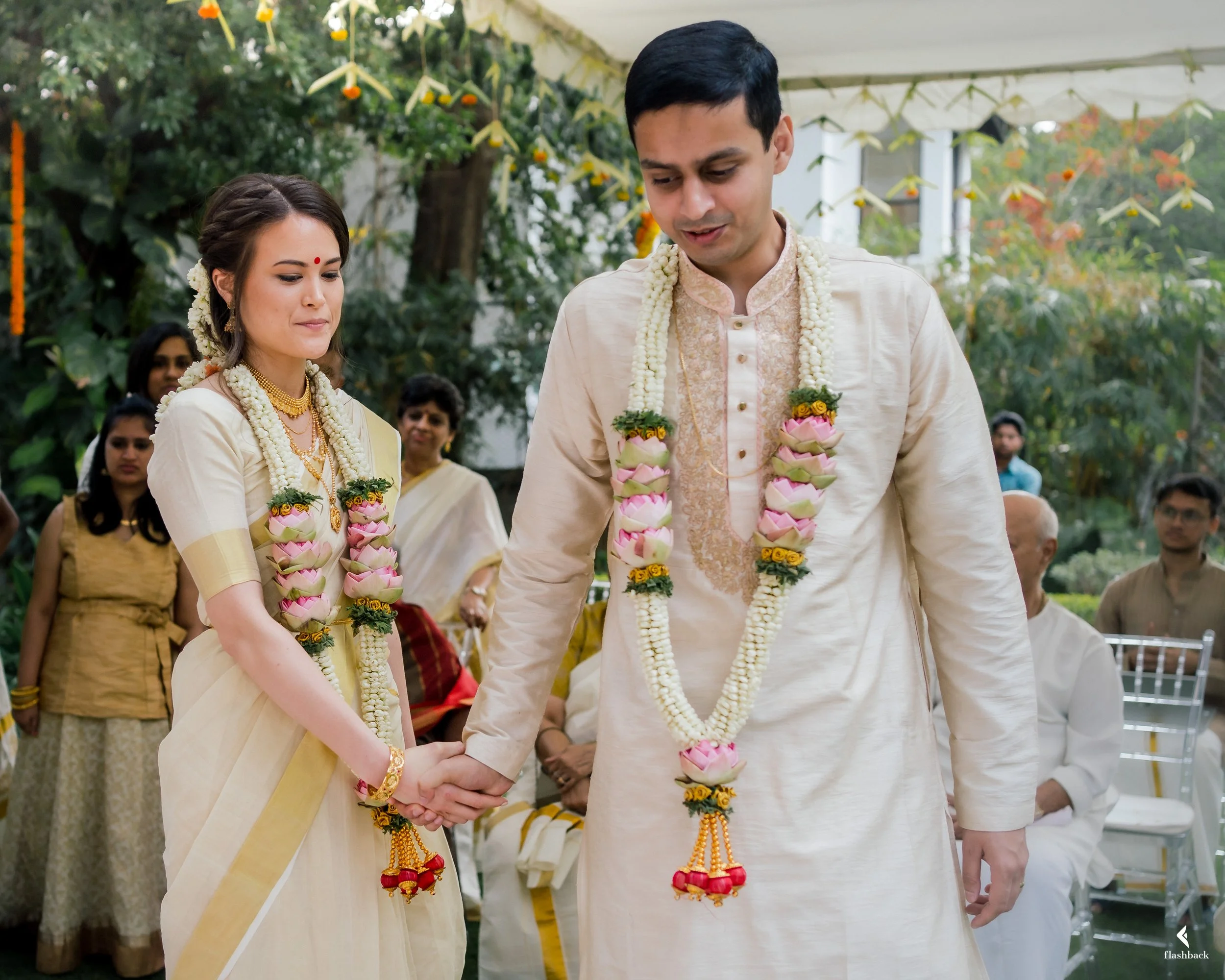 A couple in traditional Indian attire holding hands during a wedding ceremony, with guests and greenery in the background.