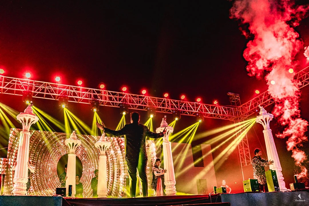 Performer on stage with arms outstretched, surrounded by colorful lights, columns, and smoke effects at a concert or event.