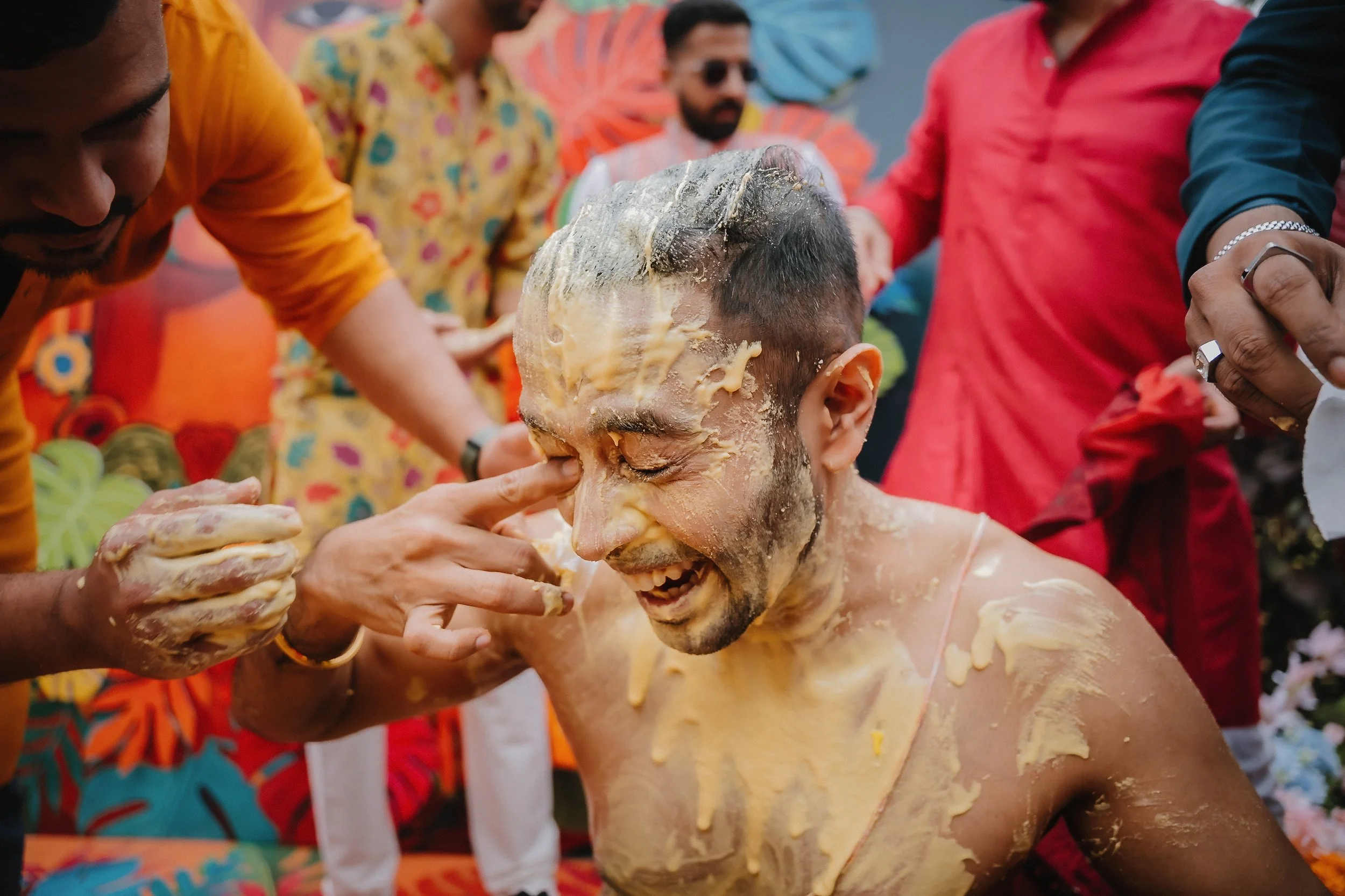 A man covered in yellowish substance, likely a mixture used in a celebration, is smiling while others pour the substance on his head during a festive outdoor event attended by people in colorful clothing.