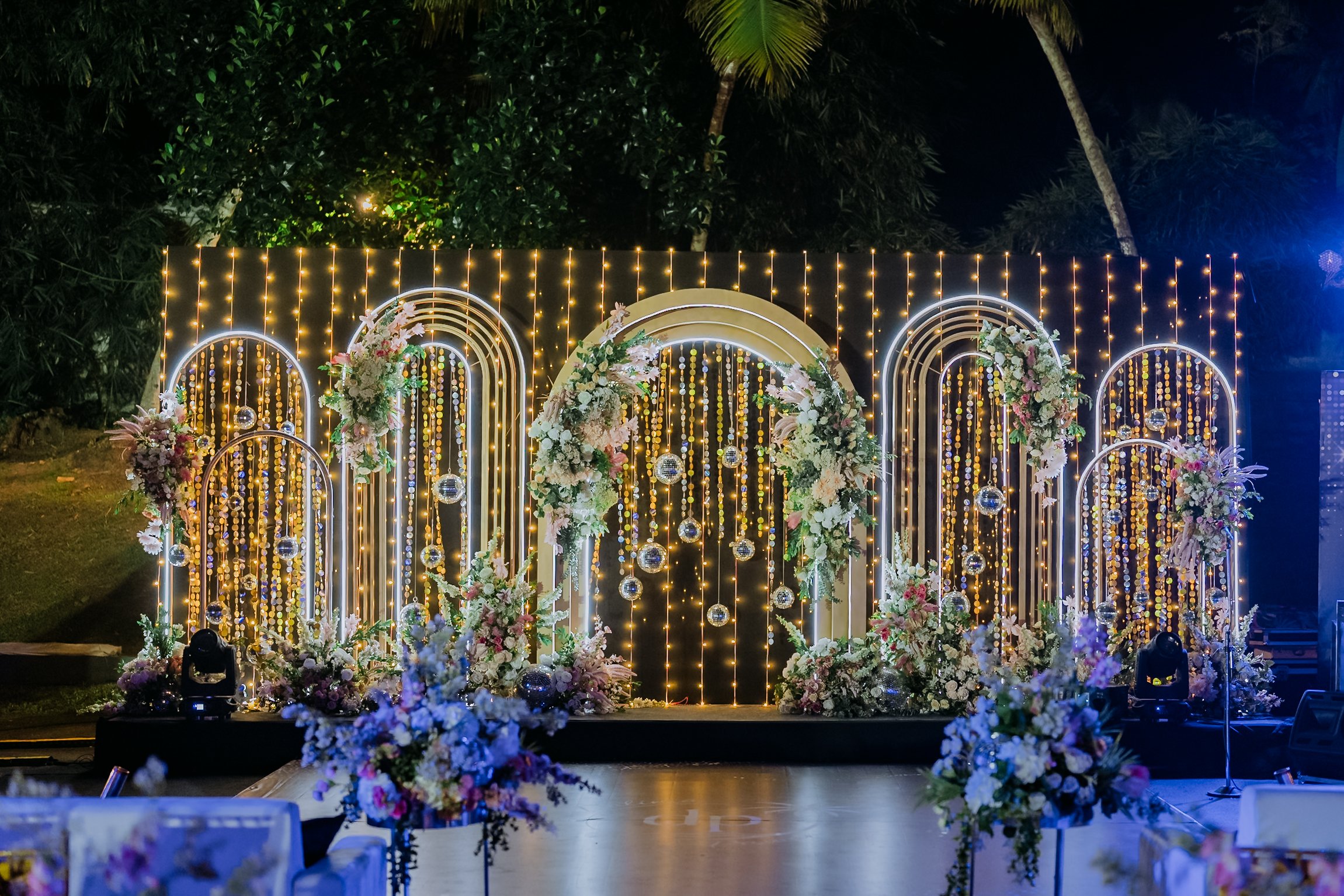 Decorative floral arch with string lights and hanging disco balls at night, likely set up for a wedding or celebration.
