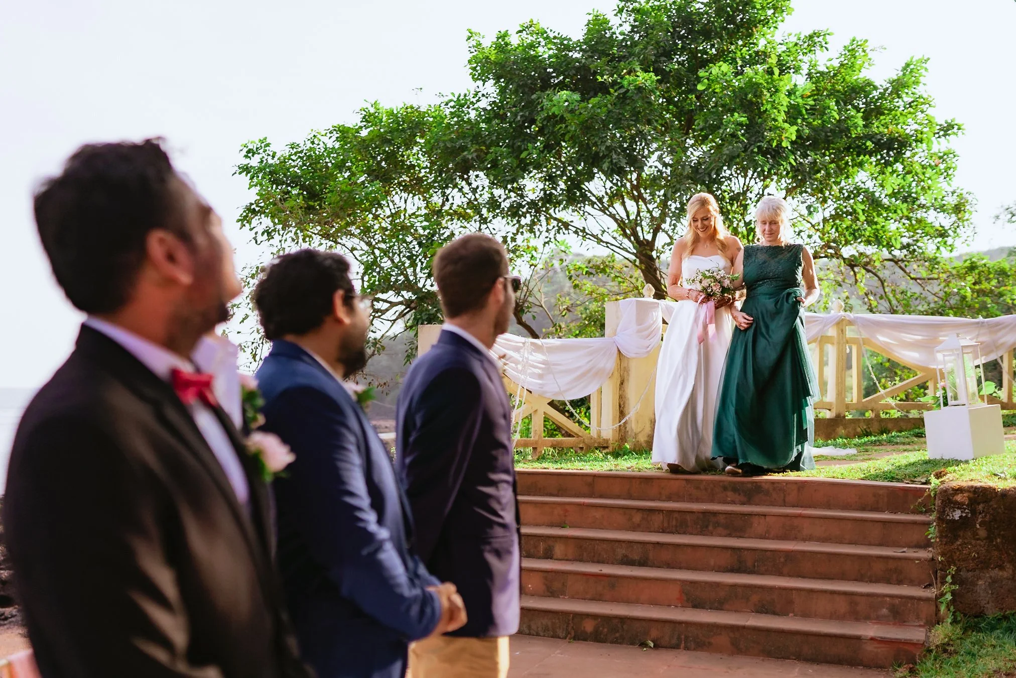 A wedding ceremony outdoors with three groomsmen in the foreground facing the stage where two women, including a bride in a white dress, are walking down the steps.