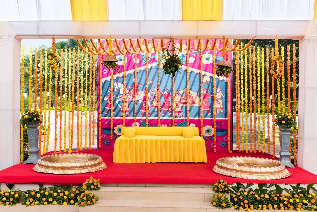 A colorful decorated stage with yellow and pink drapes, floral garlands, and hanging flower arrangements. There are two small yellow cushions on a yellow-covered bench, and two gold-colored floral pools on either side of the stage. The backdrop featu