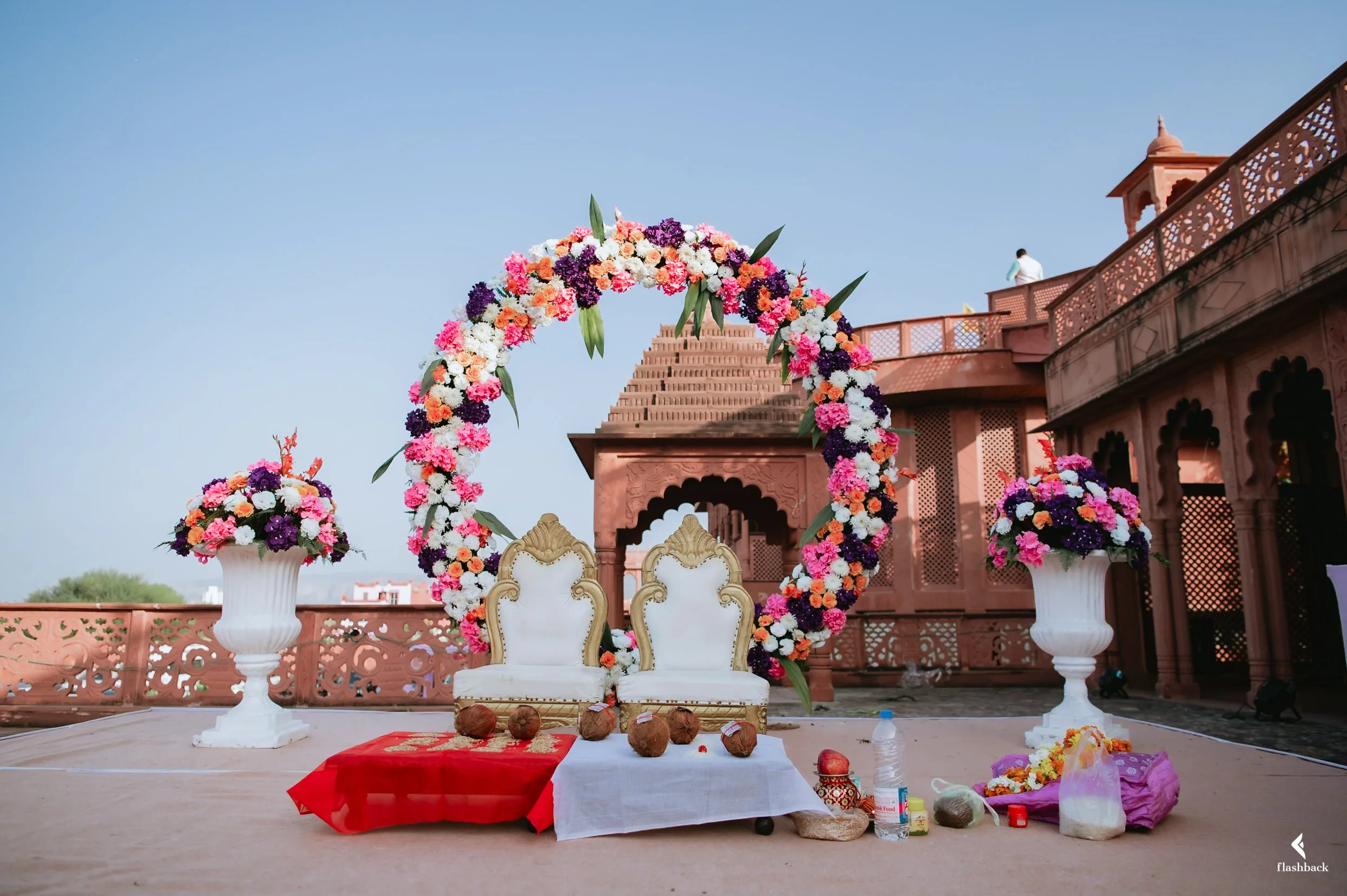 Traditional Indian wedding setup with floral arch, two ornate chairs, floral arrangements in white vases, coconut offerings, water bottle, and decorative items on the ground, set against architectural domed structure.