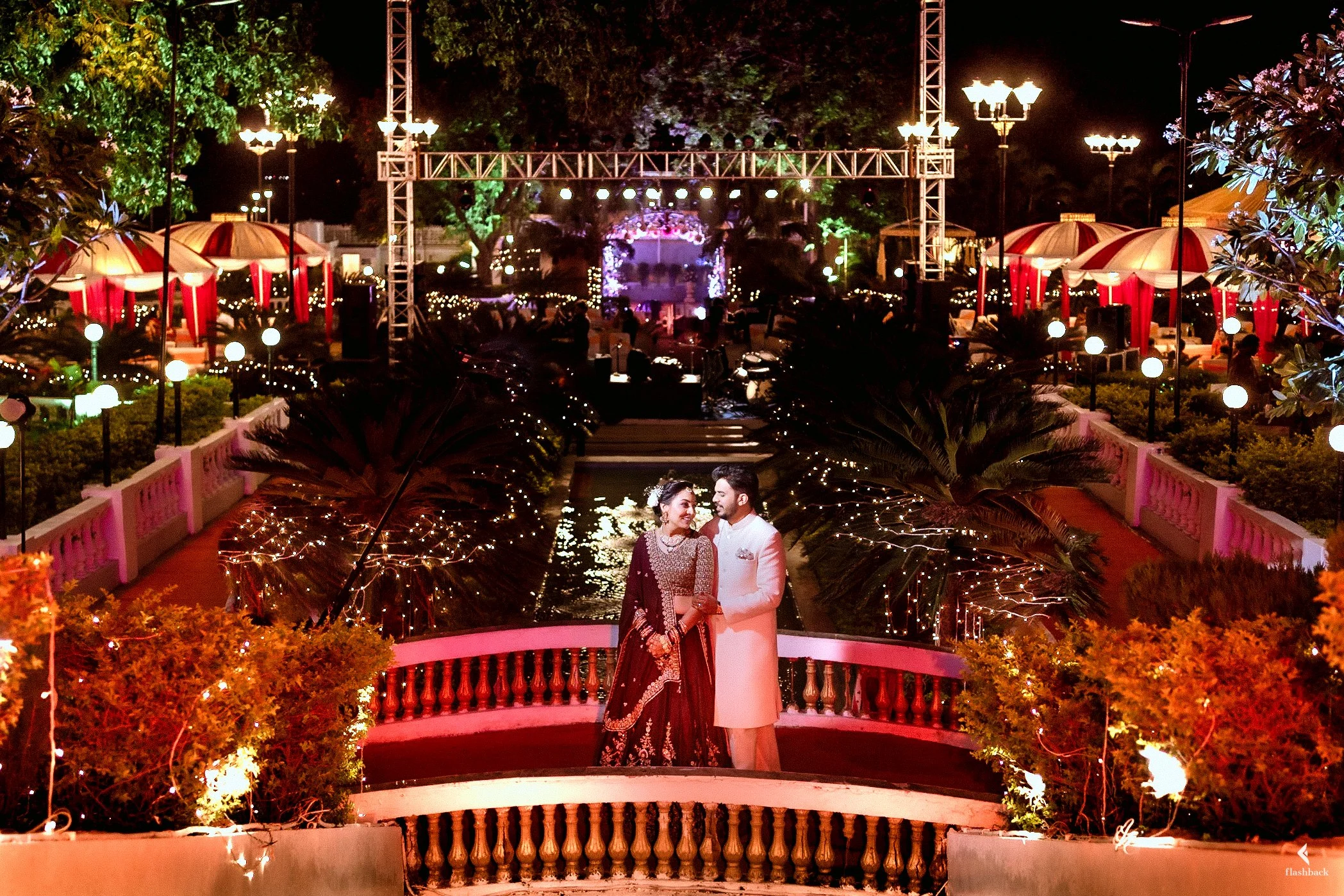 A couple dressed in traditional Indian attire stands on a decorated bridge at night, surrounded by festive lights and lush greenery, with a beautifully illuminated outdoor event in the background.