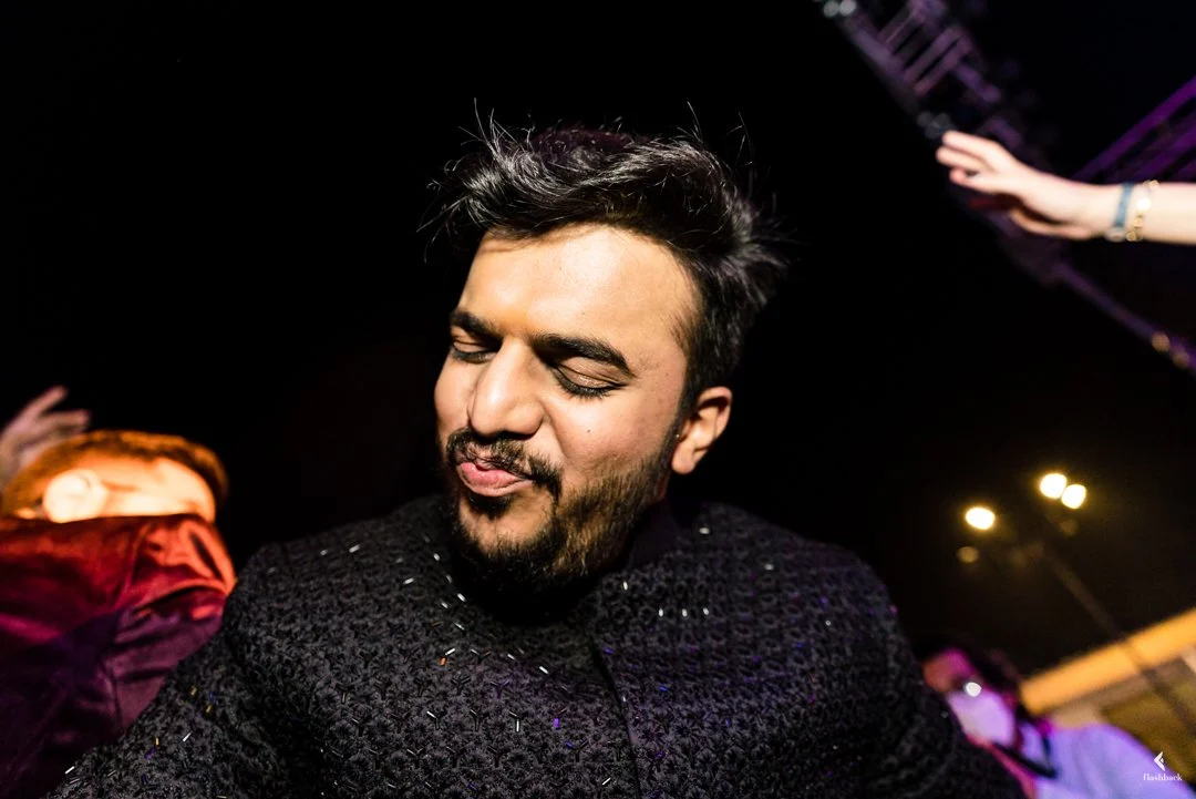 A young man with dark hair and facial hair enjoying himself at a party or club, with dark background and colorful lights.
