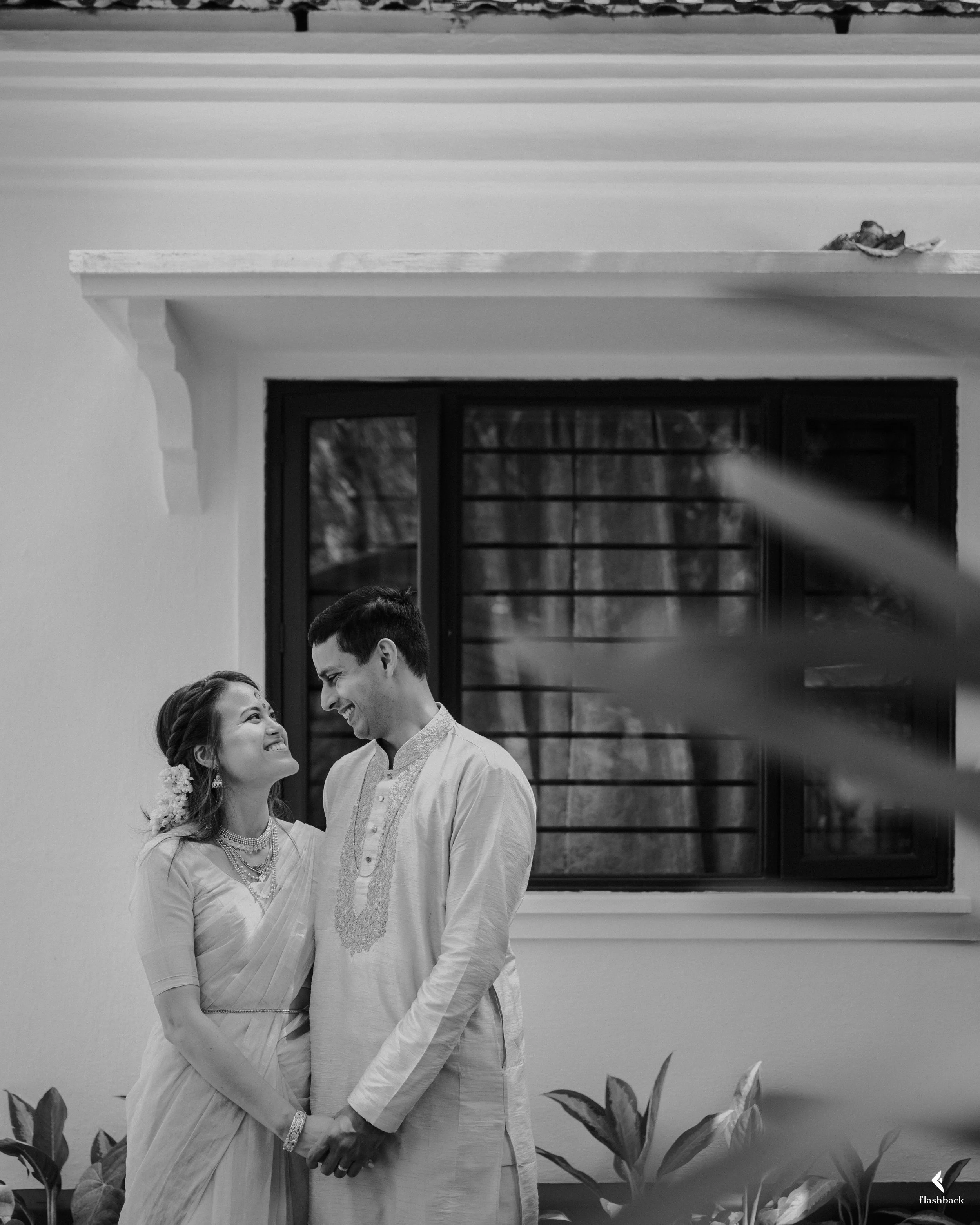 A happy couple dressed in traditional Indian attire standing close together, holding hands, and smiling at each other in front of a window with bars, with plants in the foreground.